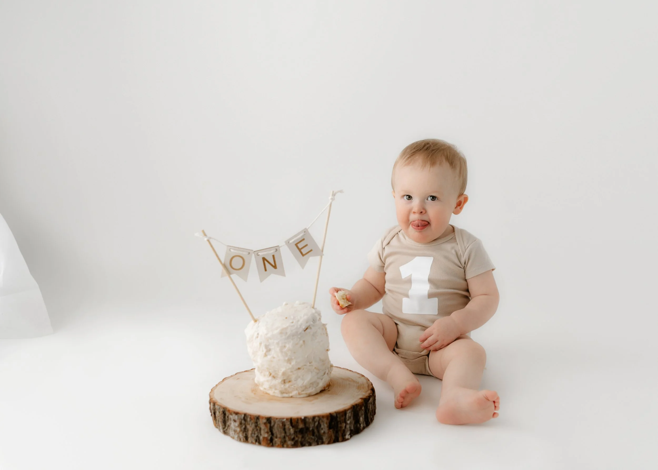 A smiling baby sitting on the floor next to a cake with a "ONE" banner, sticking out tongue, wearing a beige onesie with the number 1 on the front.