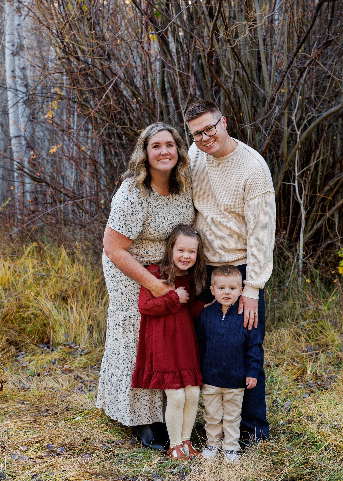 Family of five outdoors, autumn trees in background, smiling. Woman in floral dress, man in cream sweater, girl in red dress, boy in navy sweater, standing in grass.