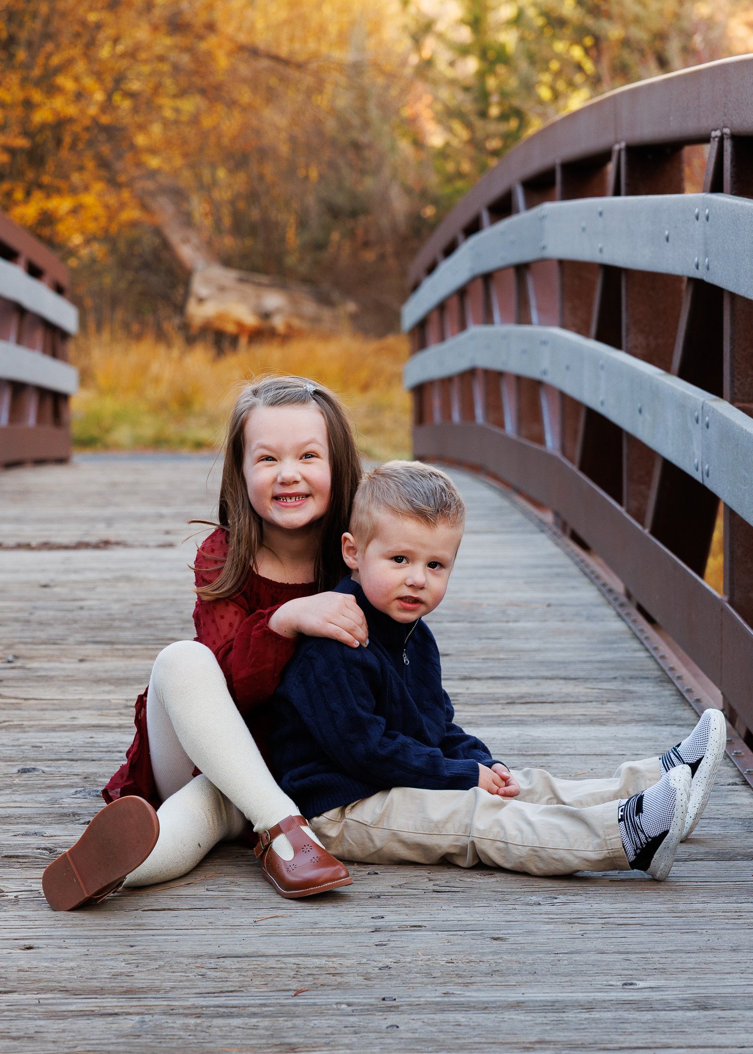 Two young children sitting on a wooden bridge in autumn, with fall foliage in the background.