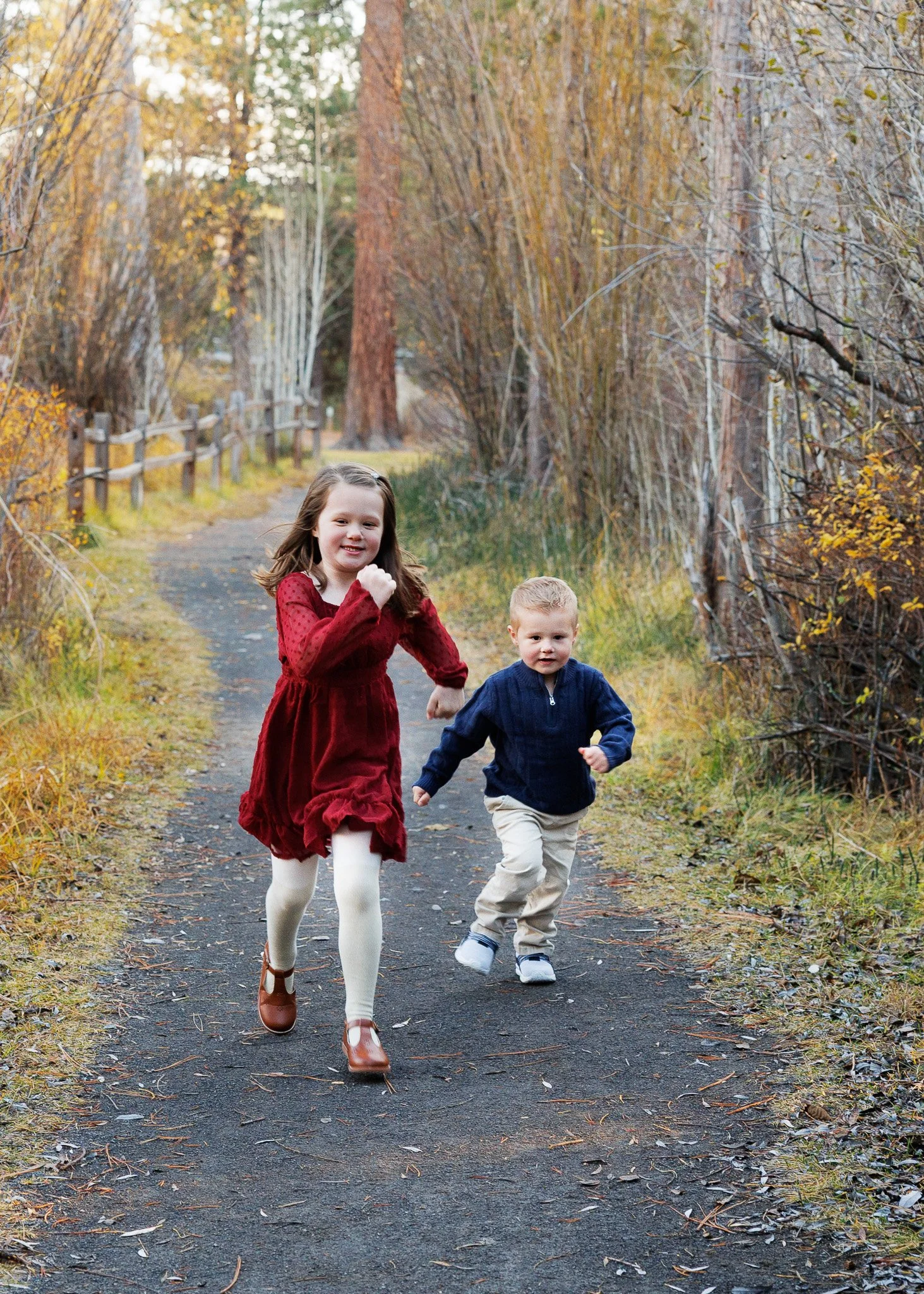 Two children, a girl in a red dress and white tights and a boy in a dark blue jacket and beige pants, running and playing on a forest trail in autumn.