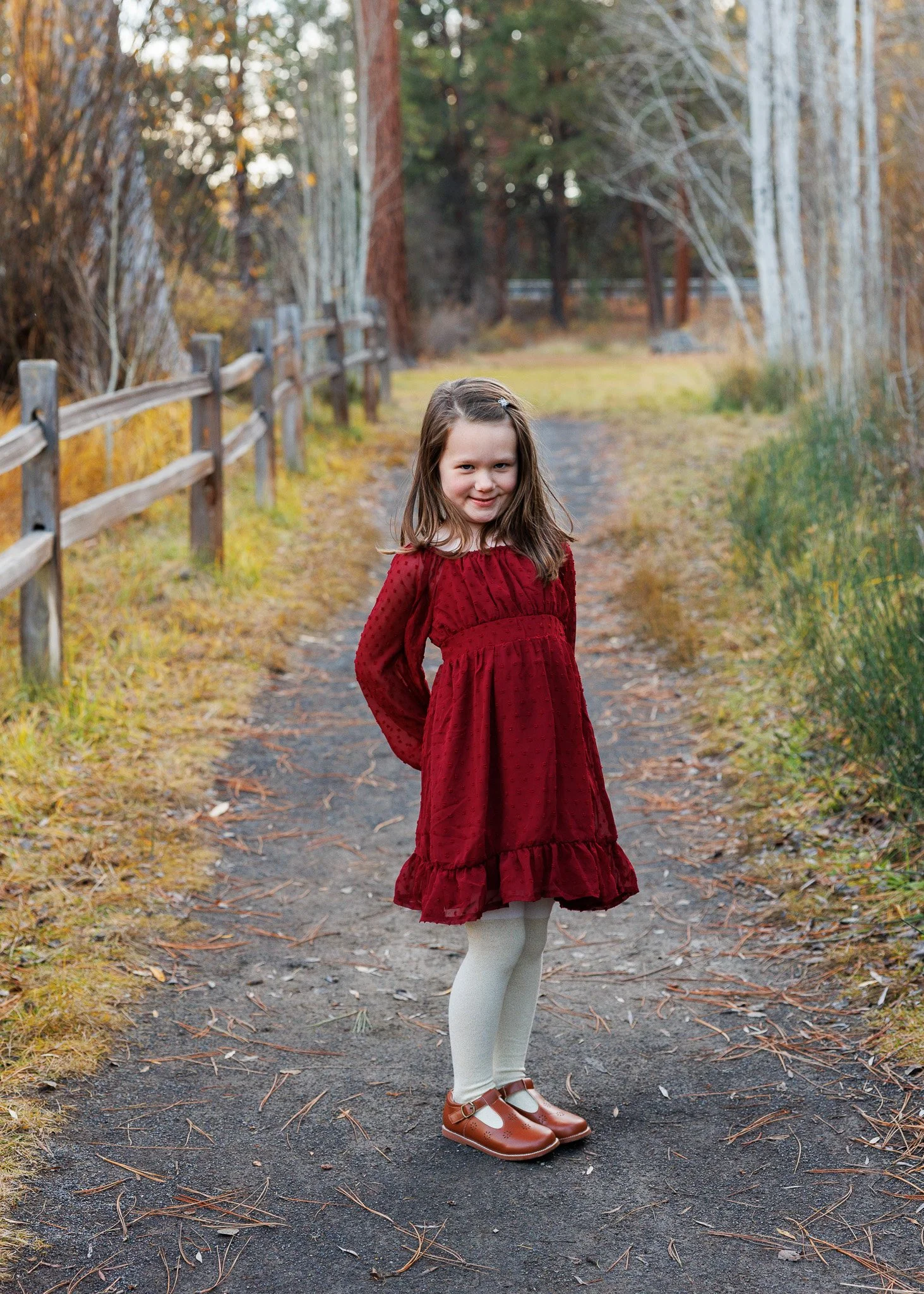 A young girl in a red dress and white tights standing on a wooded trail with trees and a wooden fence.