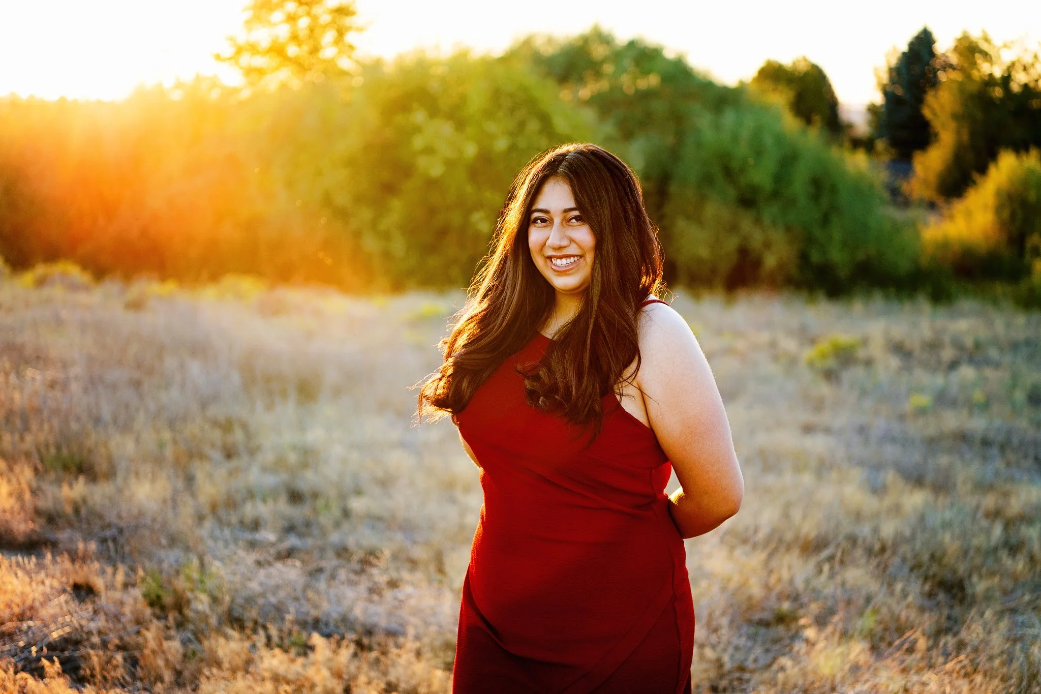 A woman with long brown hair and a red sleeveless dress, smiling outdoors during sunset in a field with trees in the background.