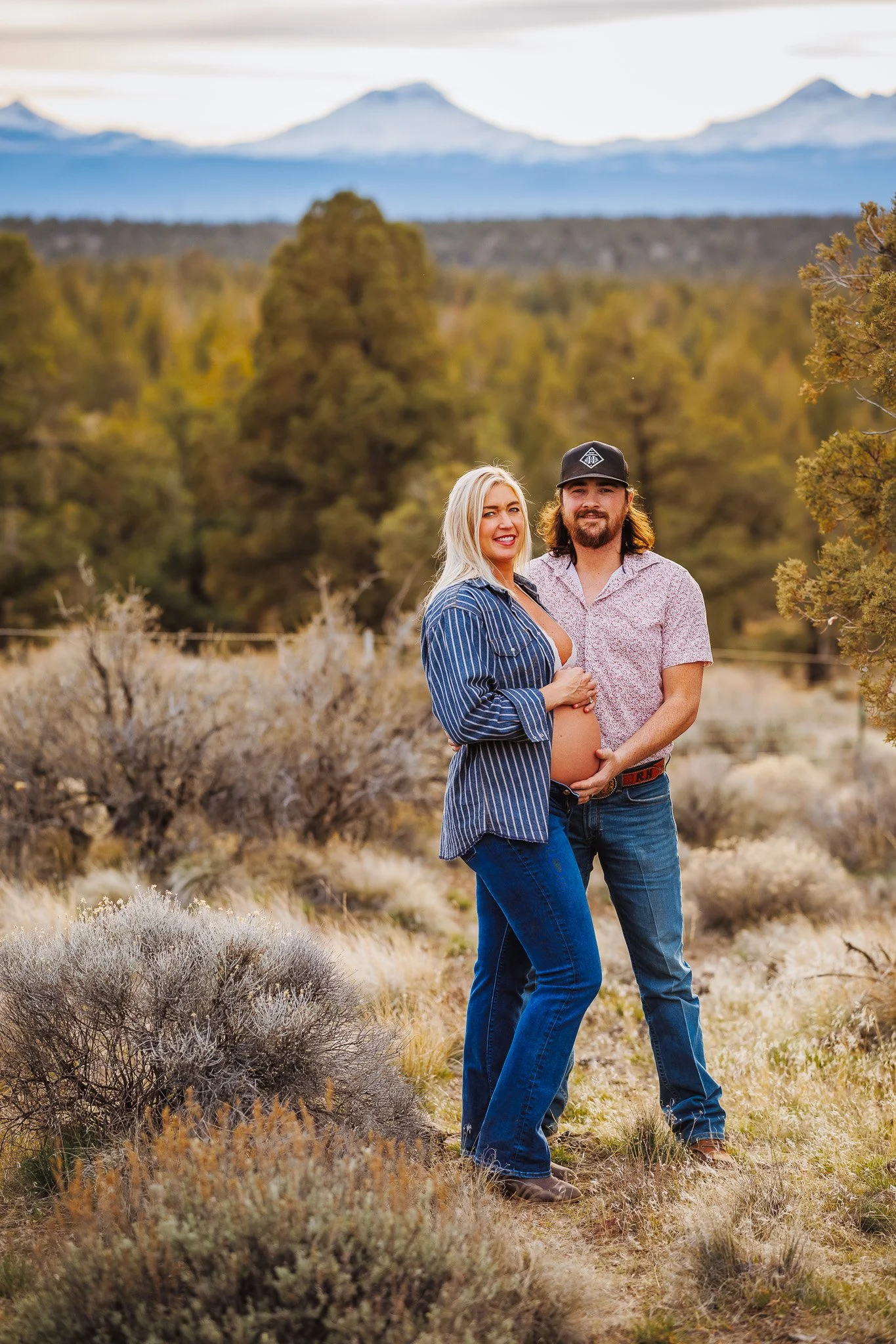 A pregnant woman and a man standing in a desert landscape with mountains in the background. The woman is smiling, wearing a striped shirt, and has her hand on her belly. The man is wearing a patterned shirt, a cap, and has his arm around the woman, b