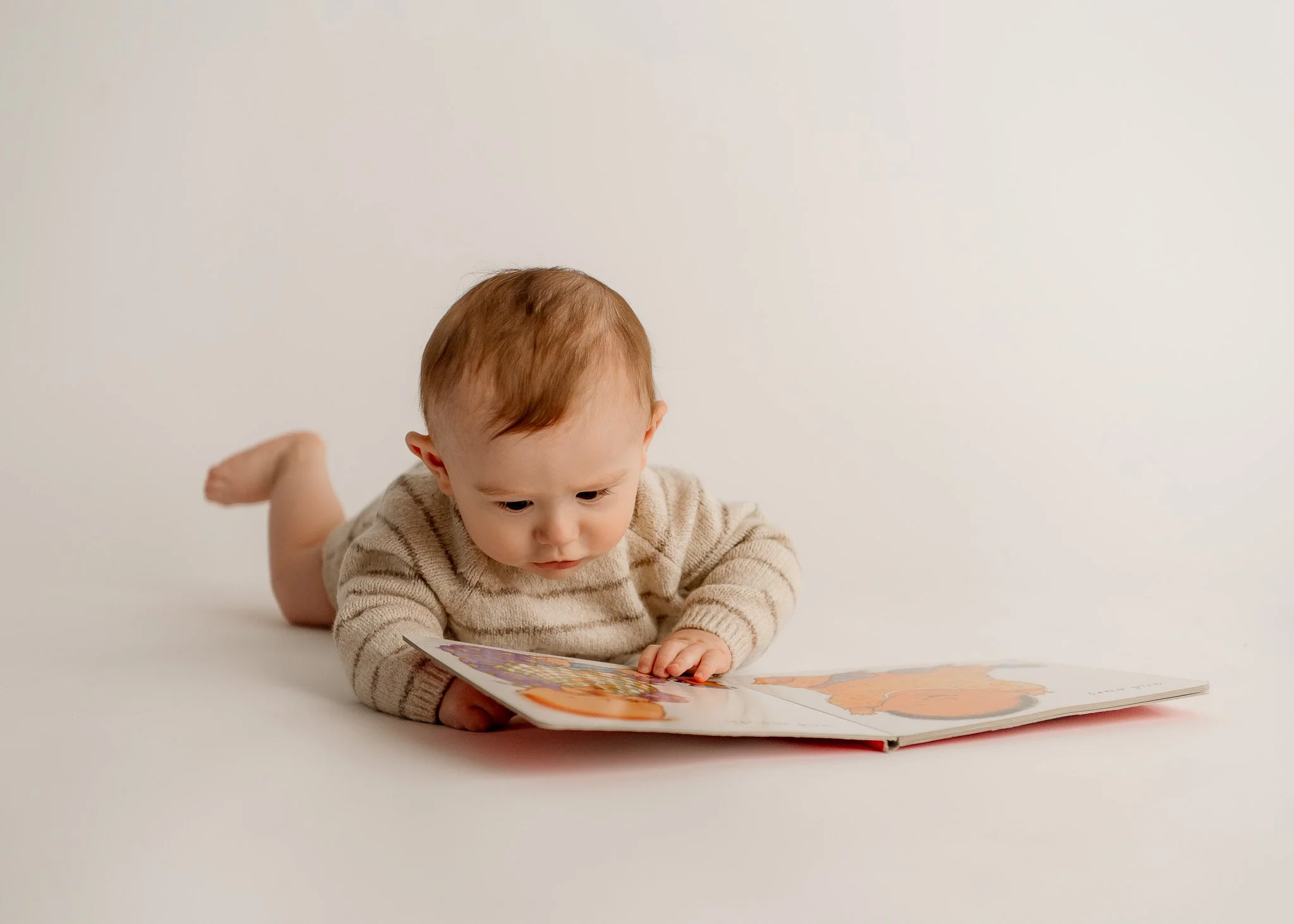 A young child with red hair and a beige striped sweater lying on their stomach, looking at a picture book on a plain white background.