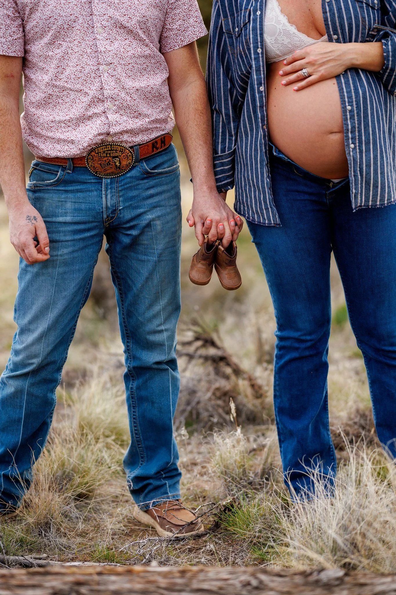 A couple standing outdoors on a grassy field, holding a small pair of baby shoes and hand in hand, with one woman showing a bare, pregnant belly.