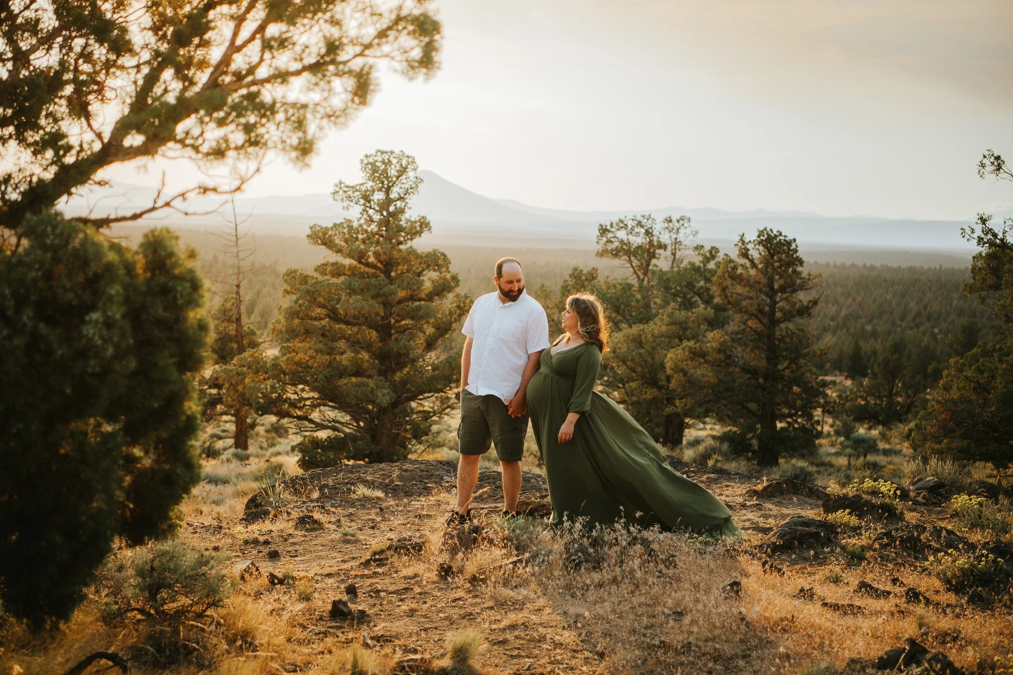 A couple holds hands on a rocky terrain in a forest clearing at sunset, with trees and mountains in the background, the woman in a long green dress and the man in a white shirt and shorts.