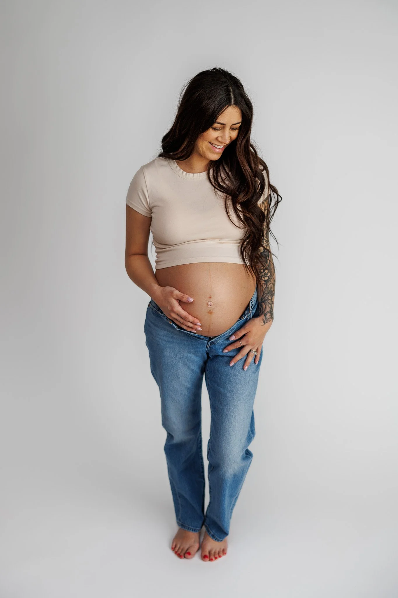 A pregnant woman with long dark hair, wearing a beige crop top and blue jeans, standing barefoot against a plain light gray background, smiling and gently touching her belly.