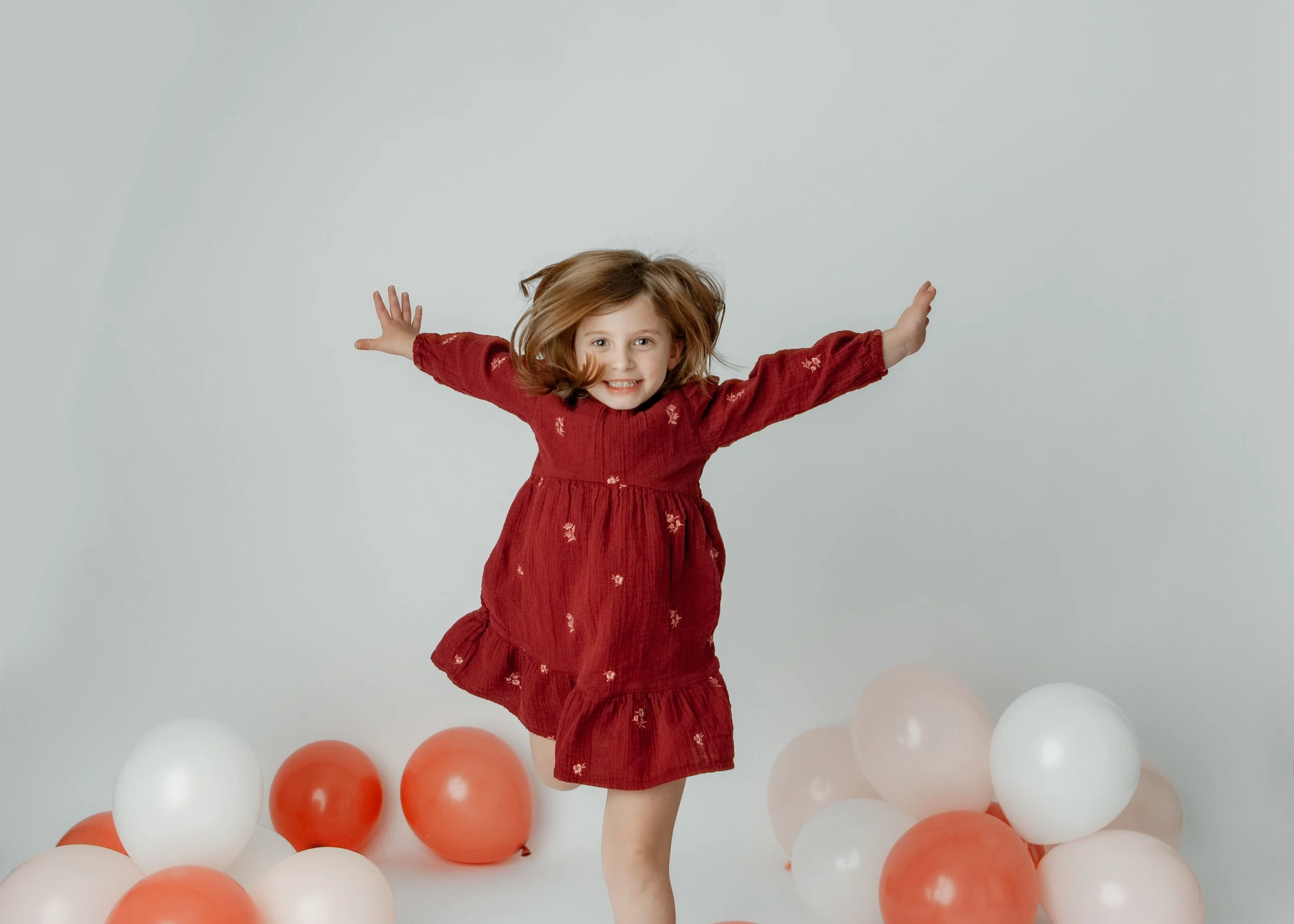 A young girl in a red dress with small floral patterns jumping and smiling among balloons of orange, white, and light pink colors on a plain light gray background.