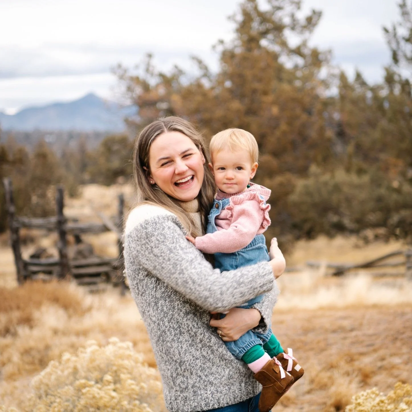 A woman with  long hair, wearing a gray sweater, smiling and holding a young girl dressed in a pink top and denim overalls in an outdoor autumn setting.
