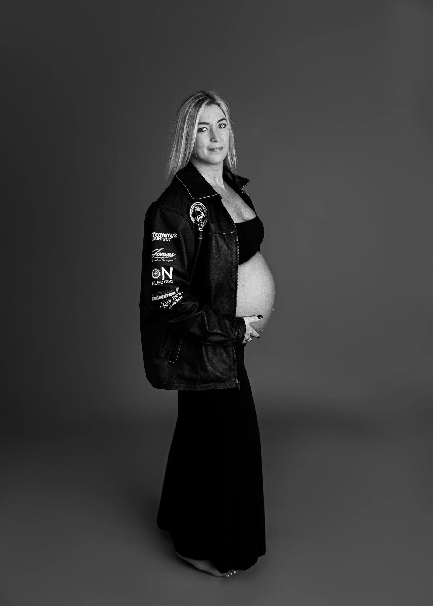 Black and white portrait of a pregnant woman with light hair, wearing a black dress and a leather jacket with various patches, standing against a plain background.