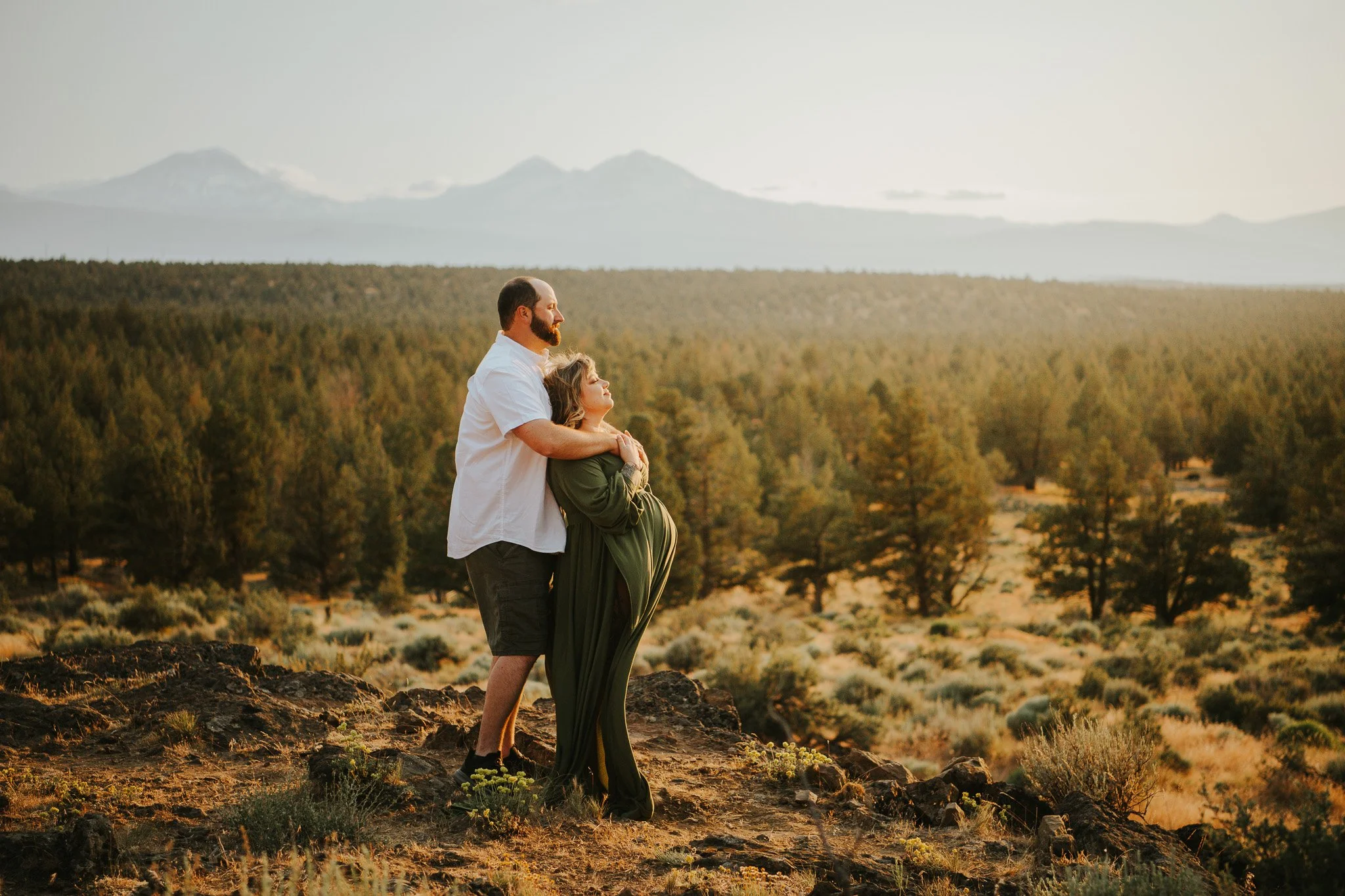 A couple embracing on a rocky hilltop with a vast forest and mountains in the background during sunset.