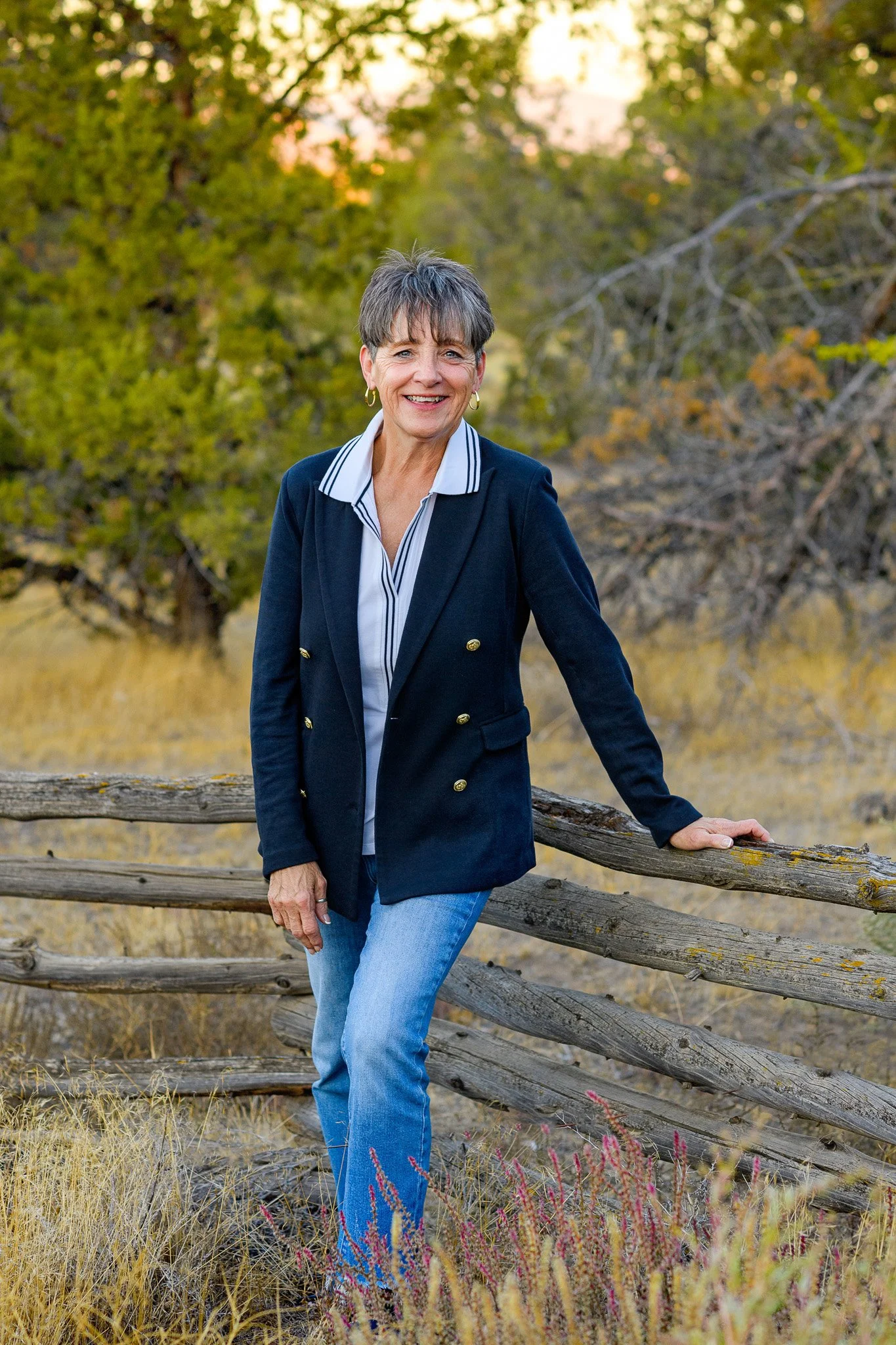A woman with short gray hair, wearing a navy blazer with gold buttons, a white collared shirt with blue stripes, and blue jeans, smiling while standing outdoors near a wooden fence with trees and yellow grass in the background.