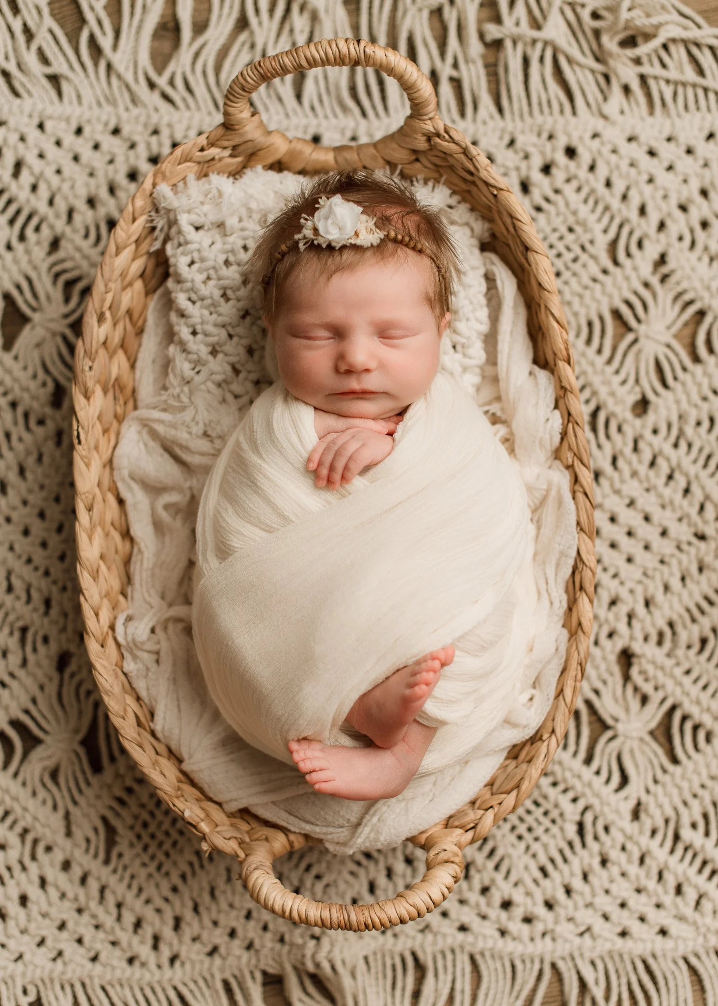 A sleeping baby wrapped in a cream blanket, lying in a woven basket on a crocheted blanket.