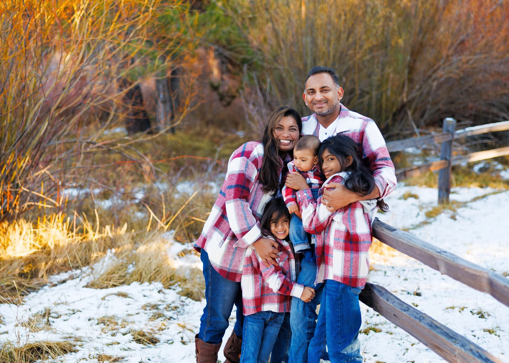 Family of five outdoors in a winter setting, dressed in plaid shirts, standing next to a wooden fence with snow on the ground and colorful autumn trees in the background.