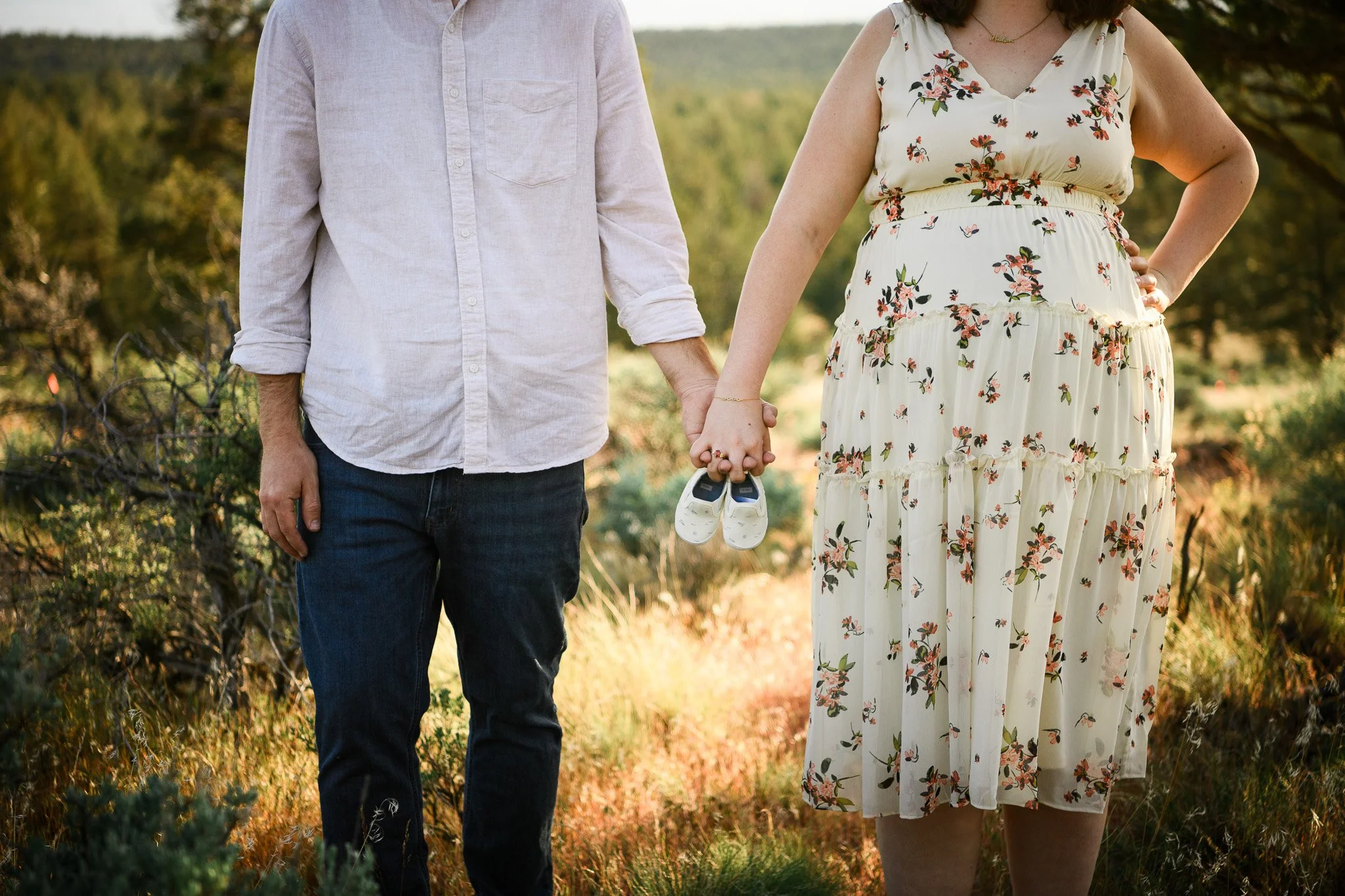 A couple standing hand in hand outdoors, holding a small pair of white baby shoes.