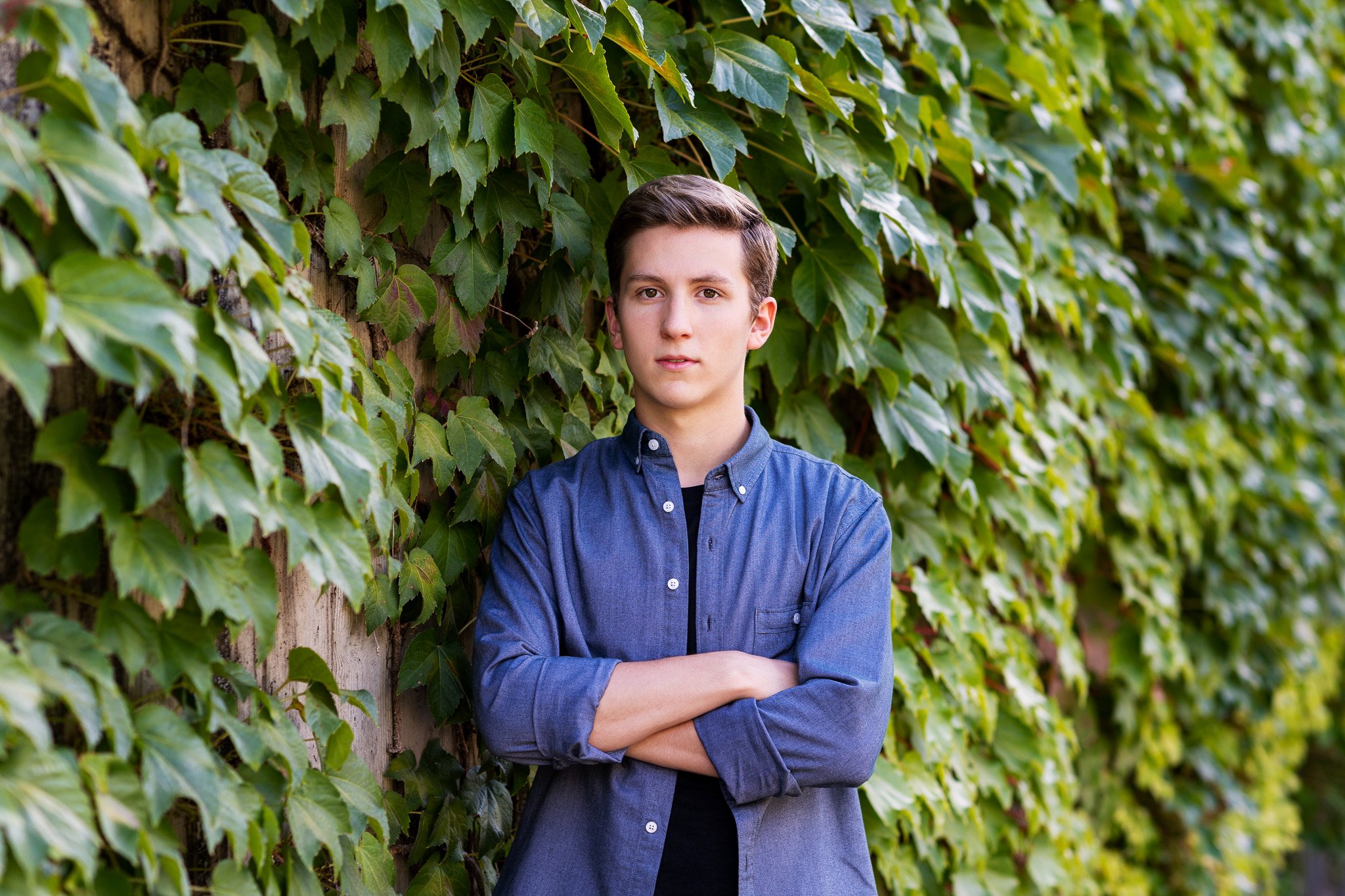 Young man in a blue button-up shirt standing with arms crossed in front of a lush green ivy-covered wall.
