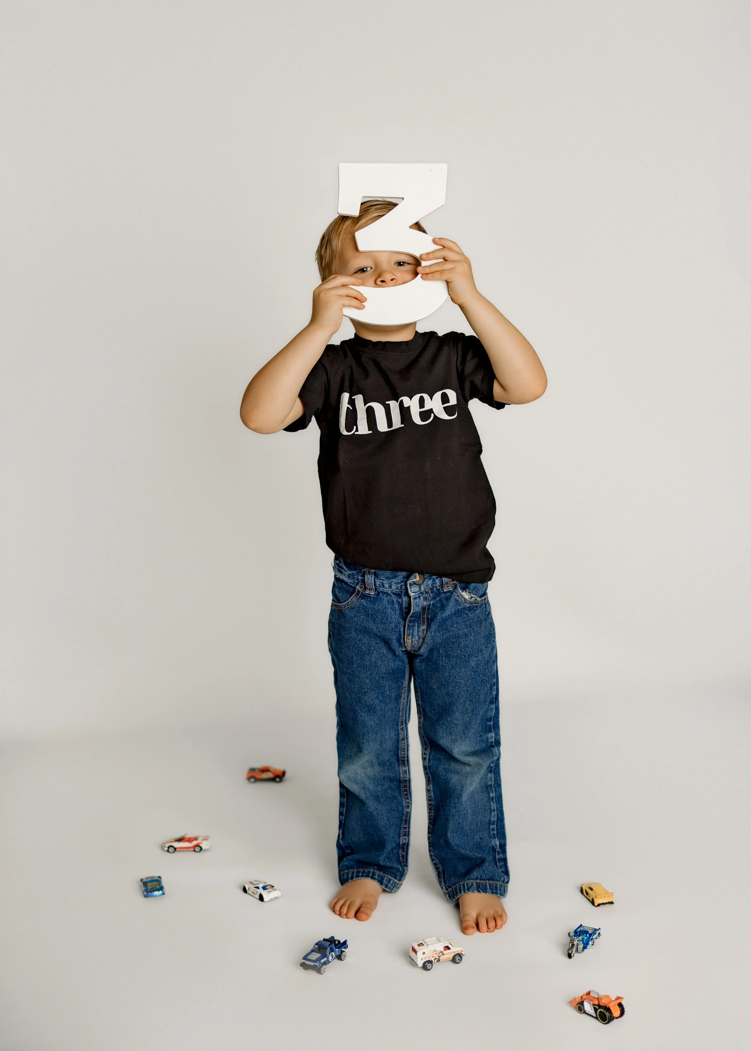 A young boy standing barefoot on a white floor, holding a large white number 3 in front of his face. He is wearing a black T-shirt with the word 'three' printed on it and blue jeans, surrounded by small toy cars placed on the floor.
