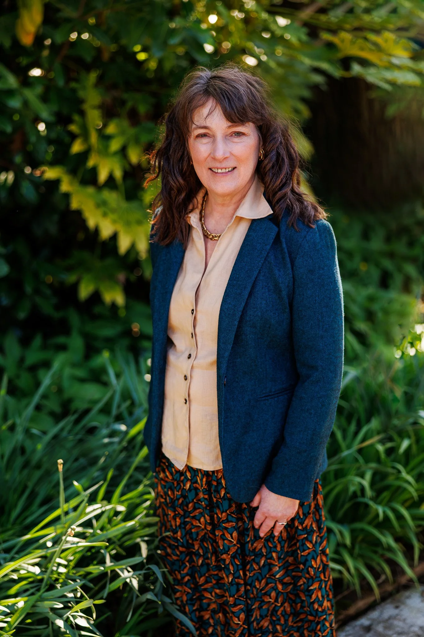A woman with shoulder-length brown hair wearing a beige blouse, a blue blazer, and patterned pants standing outdoors in front of green foliage.