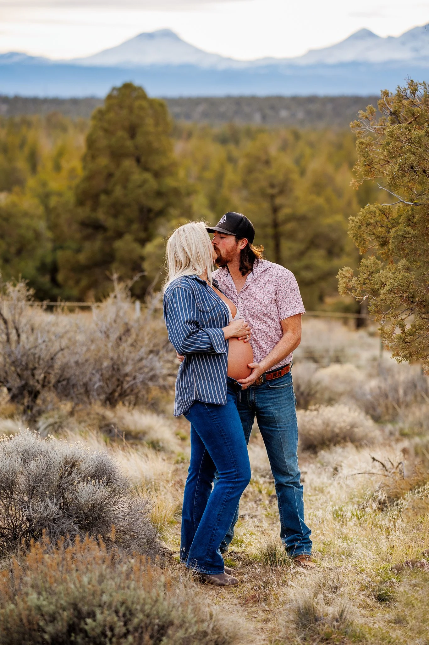 A pregnant woman and a man sharing a kiss in a natural, outdoor setting with mountains in the background.