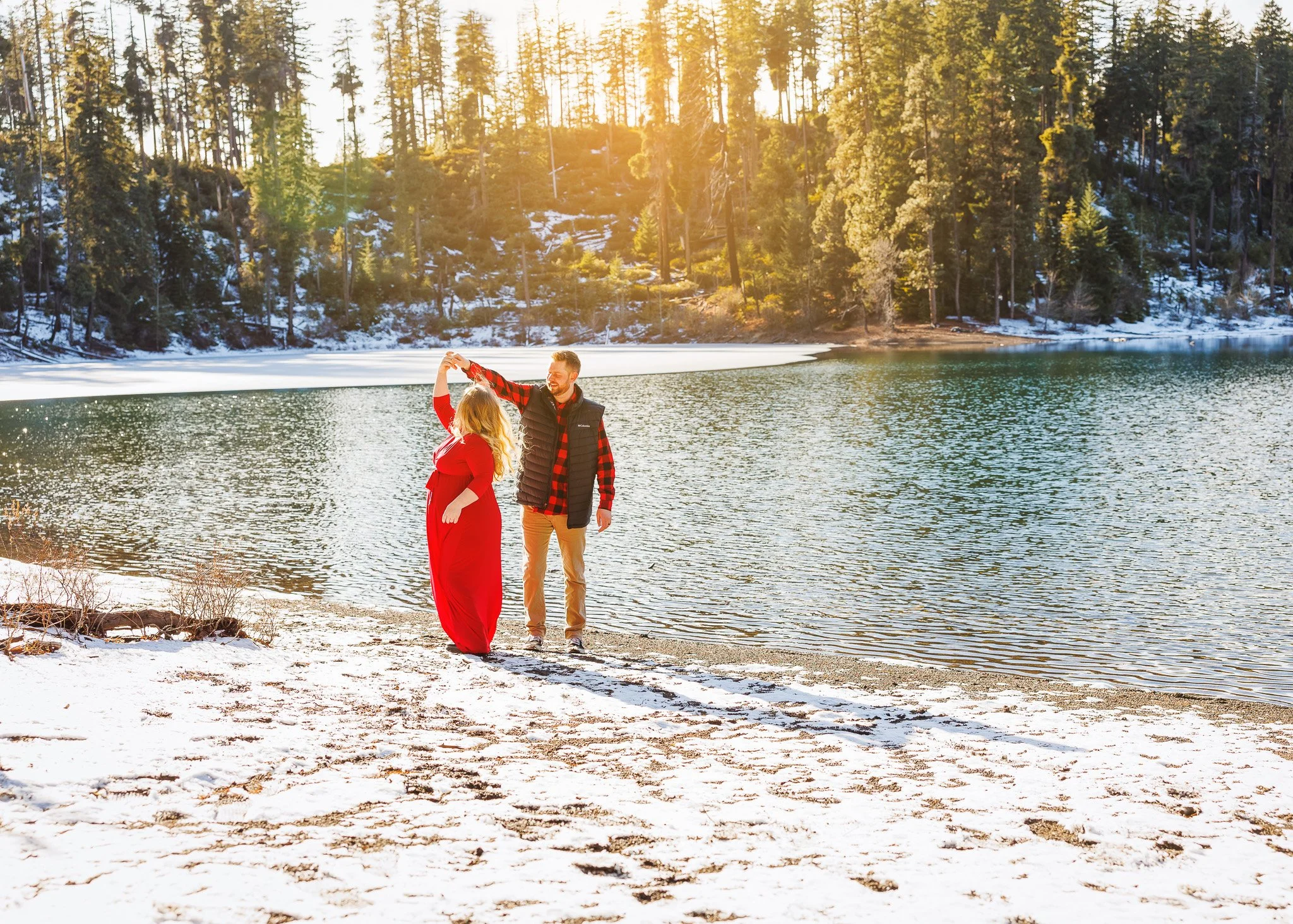 A couple dancing on the snowy shore of a lake in a forest during sunset, with trees and snow-covered ground in the background.