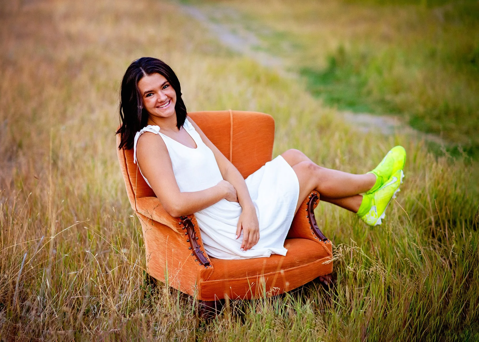 A smiling woman in a white dress and neon green sneakers relaxing on an orange armchair in a grassy field.