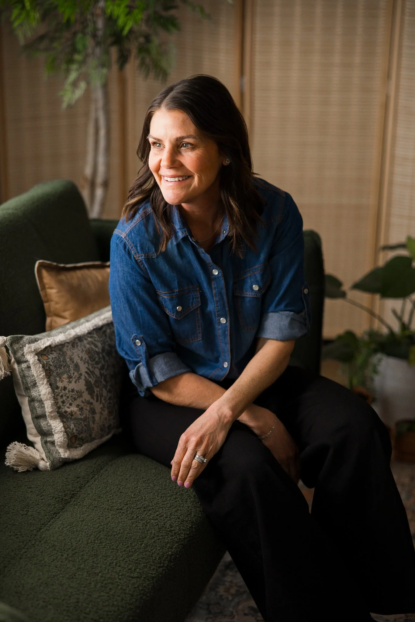 A woman in a blue denim shirt sitting on a green couch, smiling, in a cozy room with plants and wooden decor.