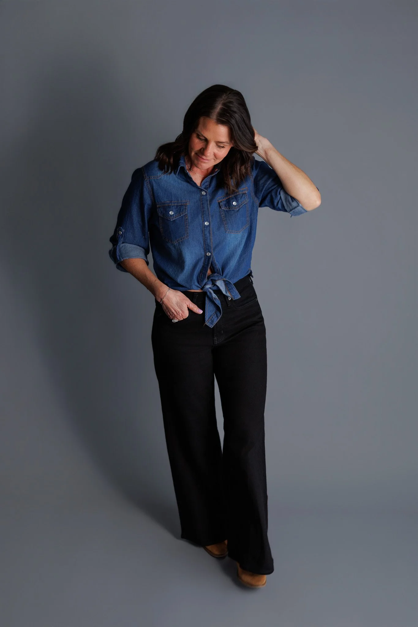 A woman standing against a gray background, wearing a blue denim button-up shirt tied at the waist, black wide-leg pants, and brown shoes. She has shoulder-length dark hair and is touching her neck with her right hand, with a relaxed expression.