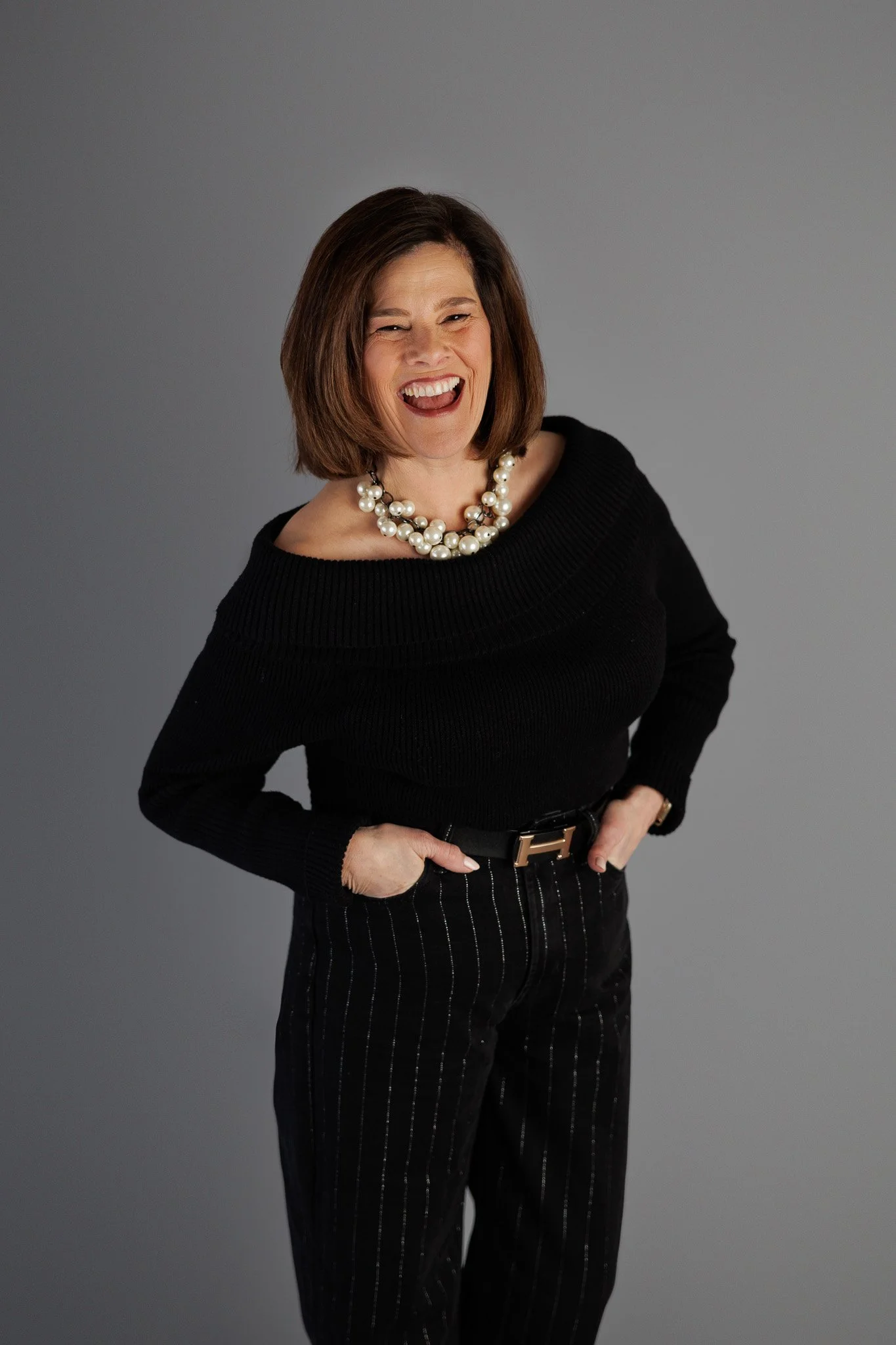 A woman with shoulder-length brown hair, wearing a black off-the-shoulder sweater, black pants with thin white pinstripes, a wide belt with a gold buckle, and a large pearl necklace, smiling and posing with her hands in her pockets against a gray bac