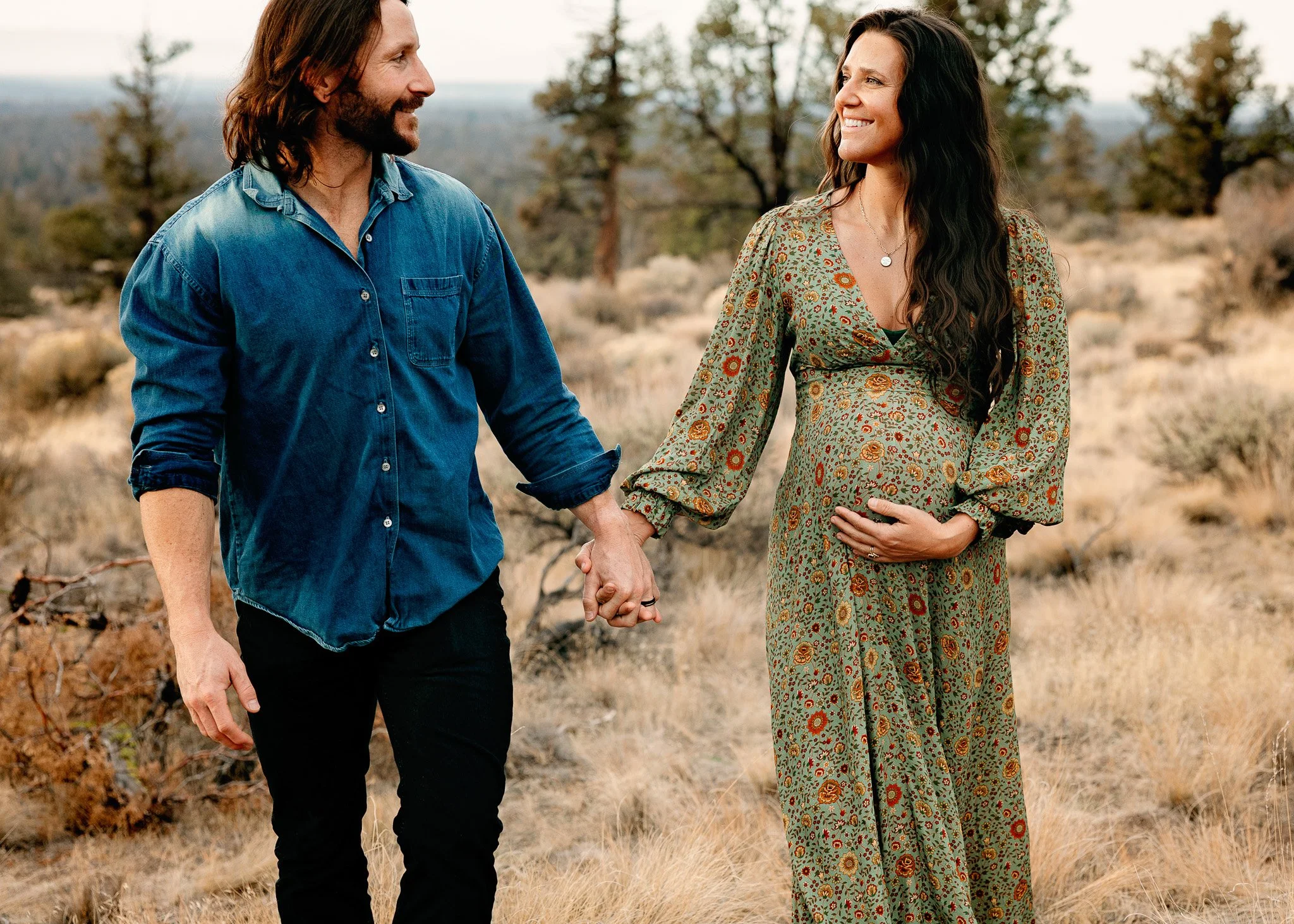 A pregnant woman and a man holding hands in a desert landscape, both smiling and looking at each other, with trees and dry grass in the background.