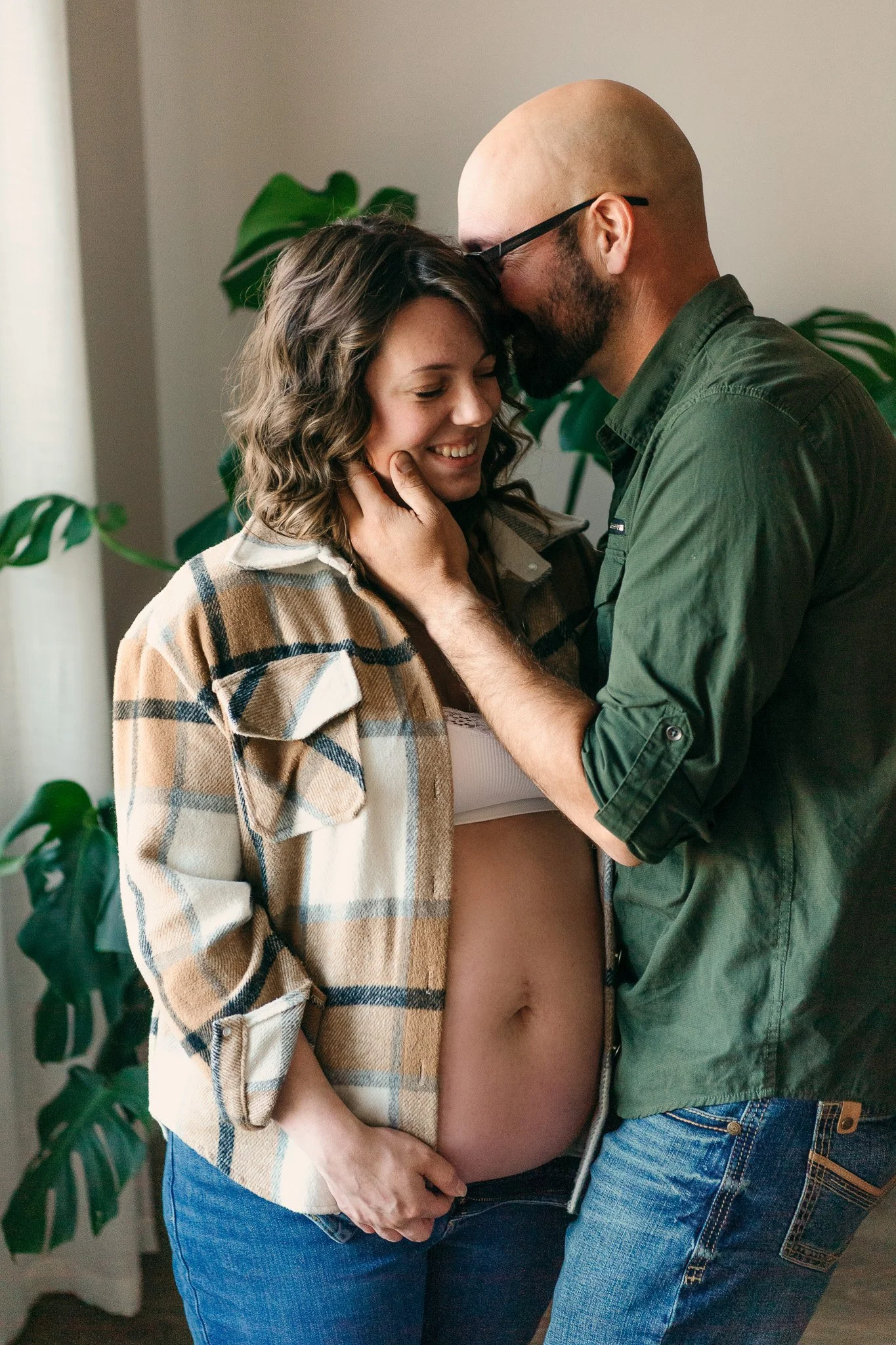 A couple sharing an intimate moment, with the woman showing her pregnant belly, smiling and touching her chin, while the man gently holds her face and kisses her forehead inside a cozy room with plants.
