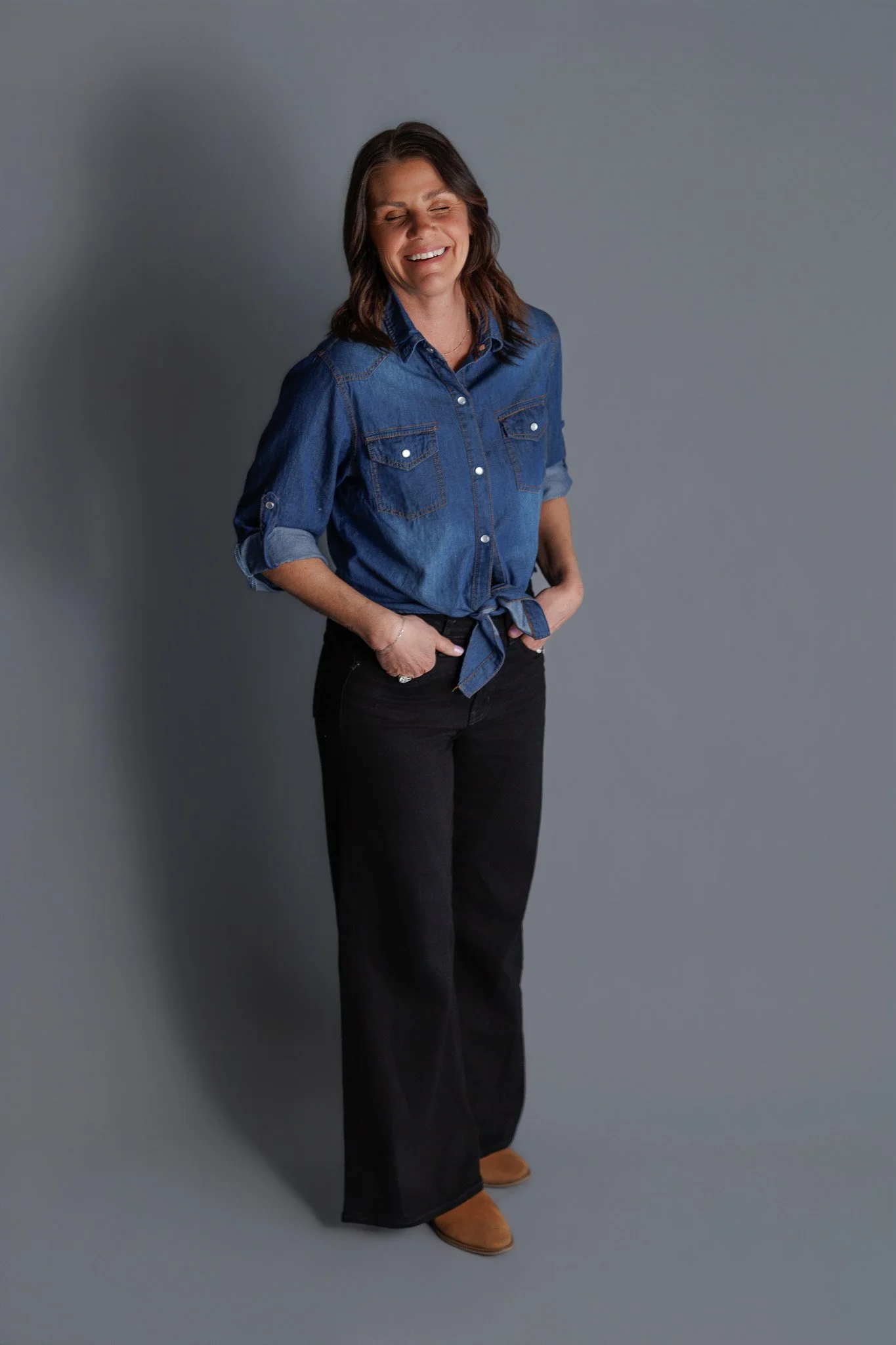 A woman standing with her eyes closed, wearing a denim shirt with rolled-up sleeves and black pants, smiling.