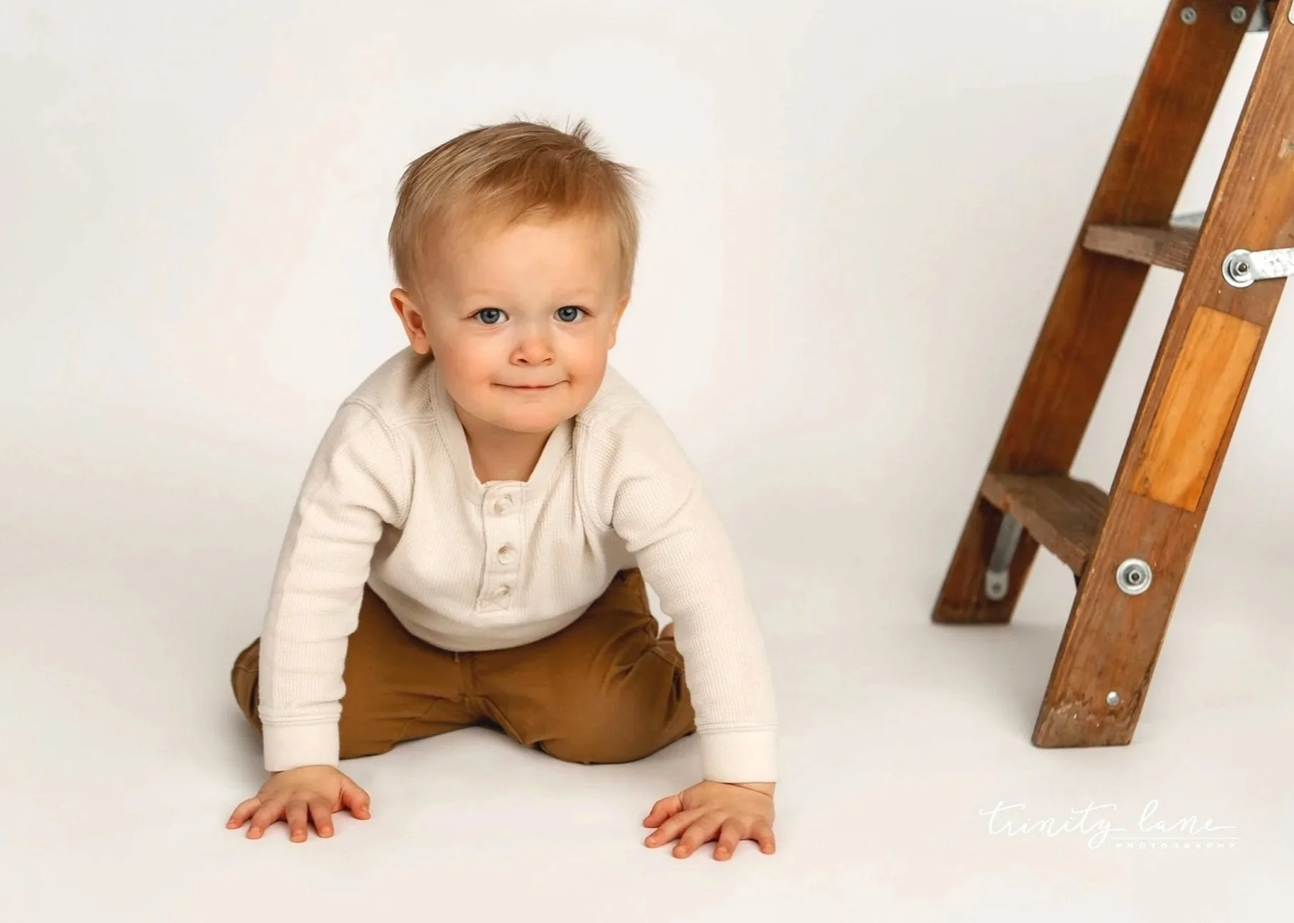 A young boy with blonde hair and blue eyes crawling on a white floor next to a wooden ladder.