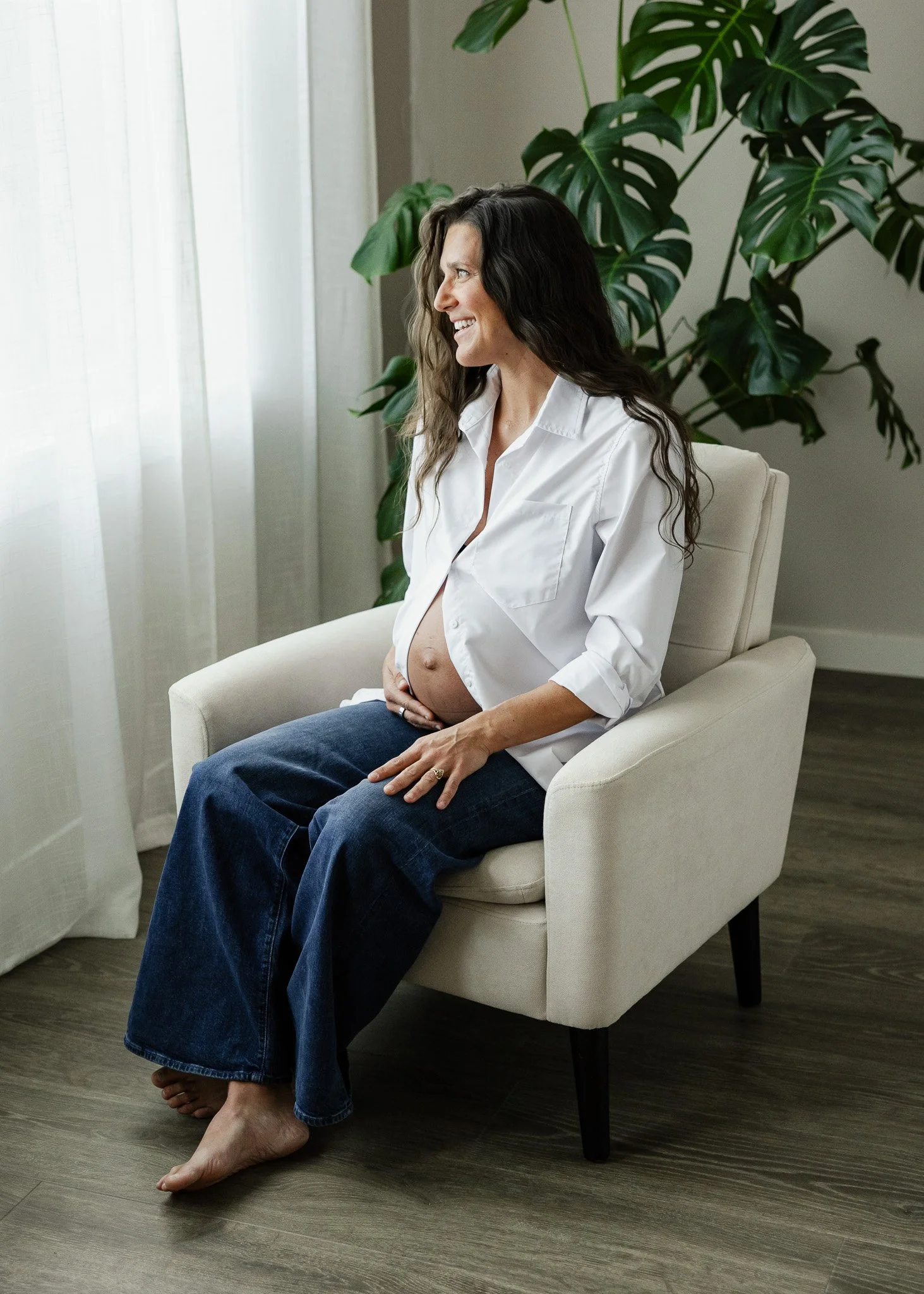 A pregnant woman sitting on a white armchair, smiling and holding her belly, indoors near a large plant and sheer curtains.