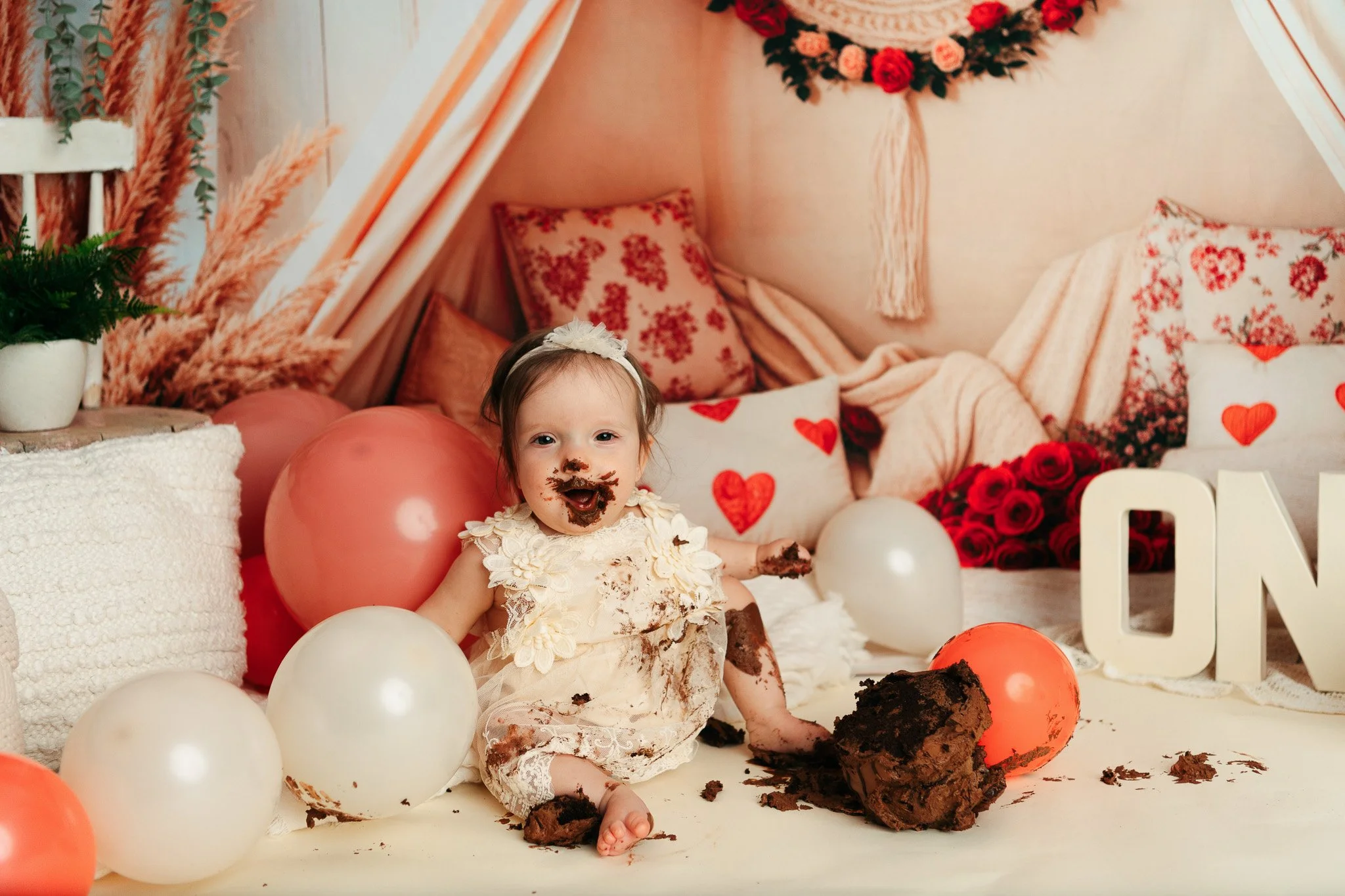 A young girl with chocolate cake on her face and clothes, sitting among balloons and cake remnants, in a decorated room with pink and red accents, heart-themed pillows, and a bouquet of red roses.