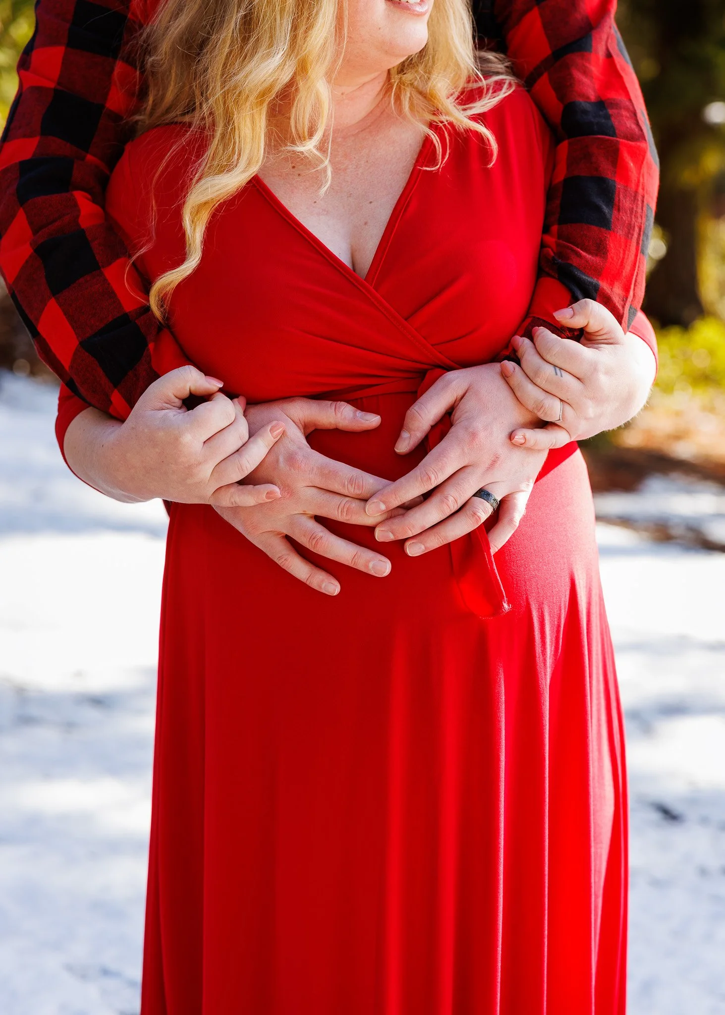 Close-up of a woman in a red dress with a person behind her, both holding their hands on her belly, outdoors in a snowy setting with blurred trees in the background.