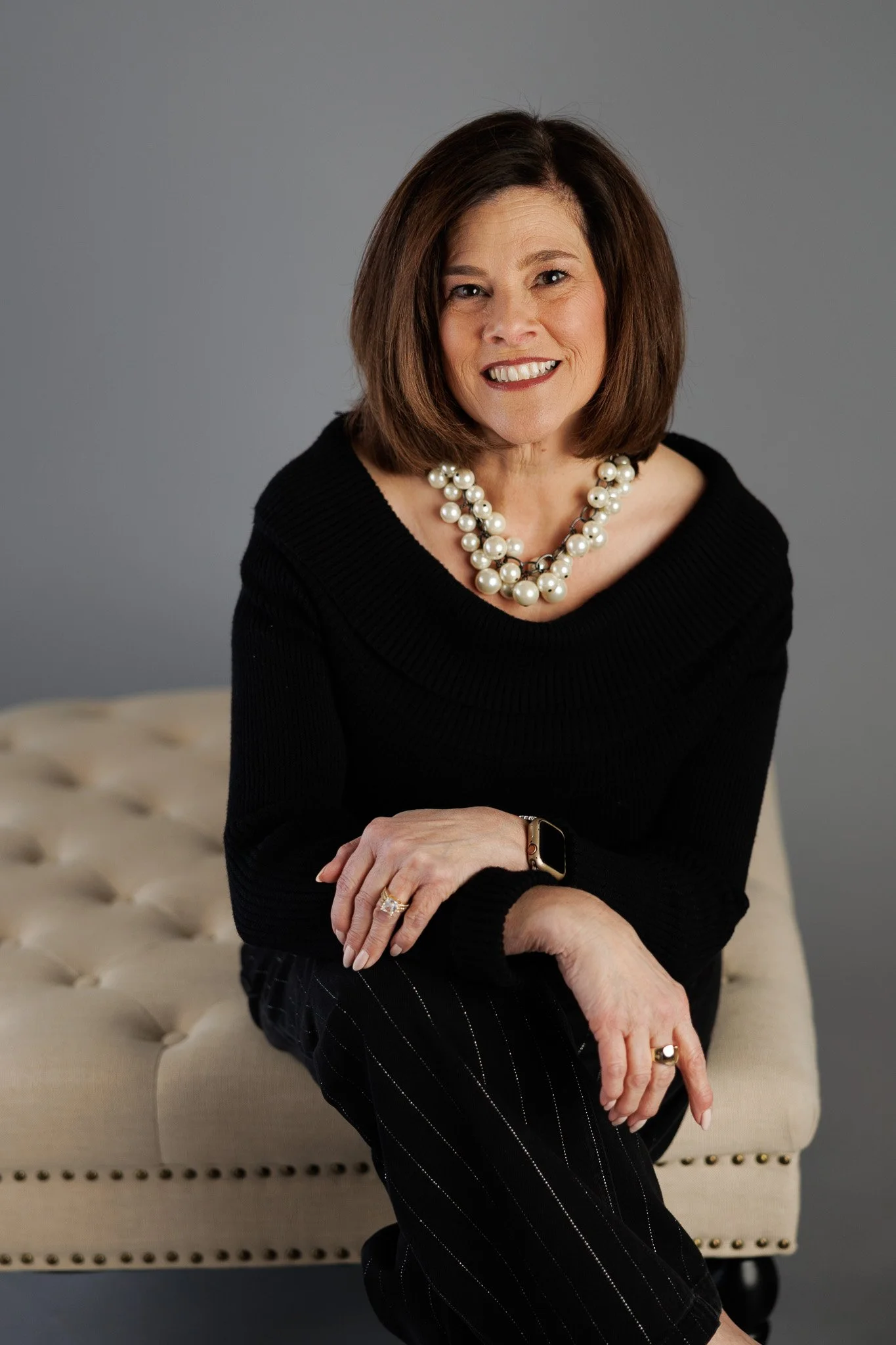 A woman with shoulder-length brown hair, wearing a black sweater and pearl necklace, sitting on a beige tufted bench against a gray background.