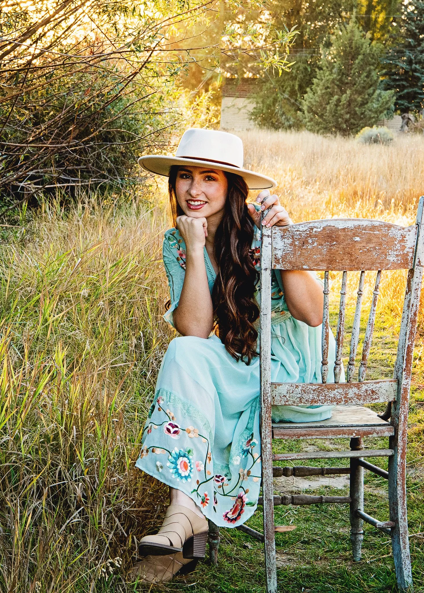 A young woman with long brown hair, wearing a wide-brimmed hat, a floral embroidered dress, and beige boots, sitting outdoors on a rustic wooden chair in a grassy field with trees in the background during golden hour sunlight.