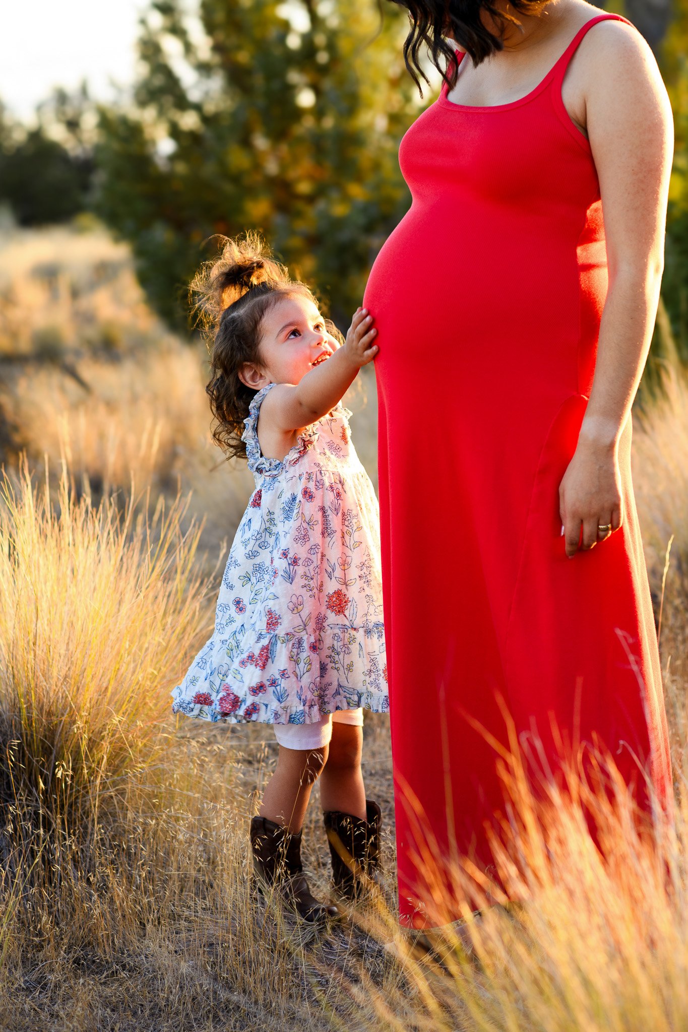 A pregnant woman in a red dress standing outdoors in a field during sunset, with a young girl touching her pregnant belly and looking up at her.