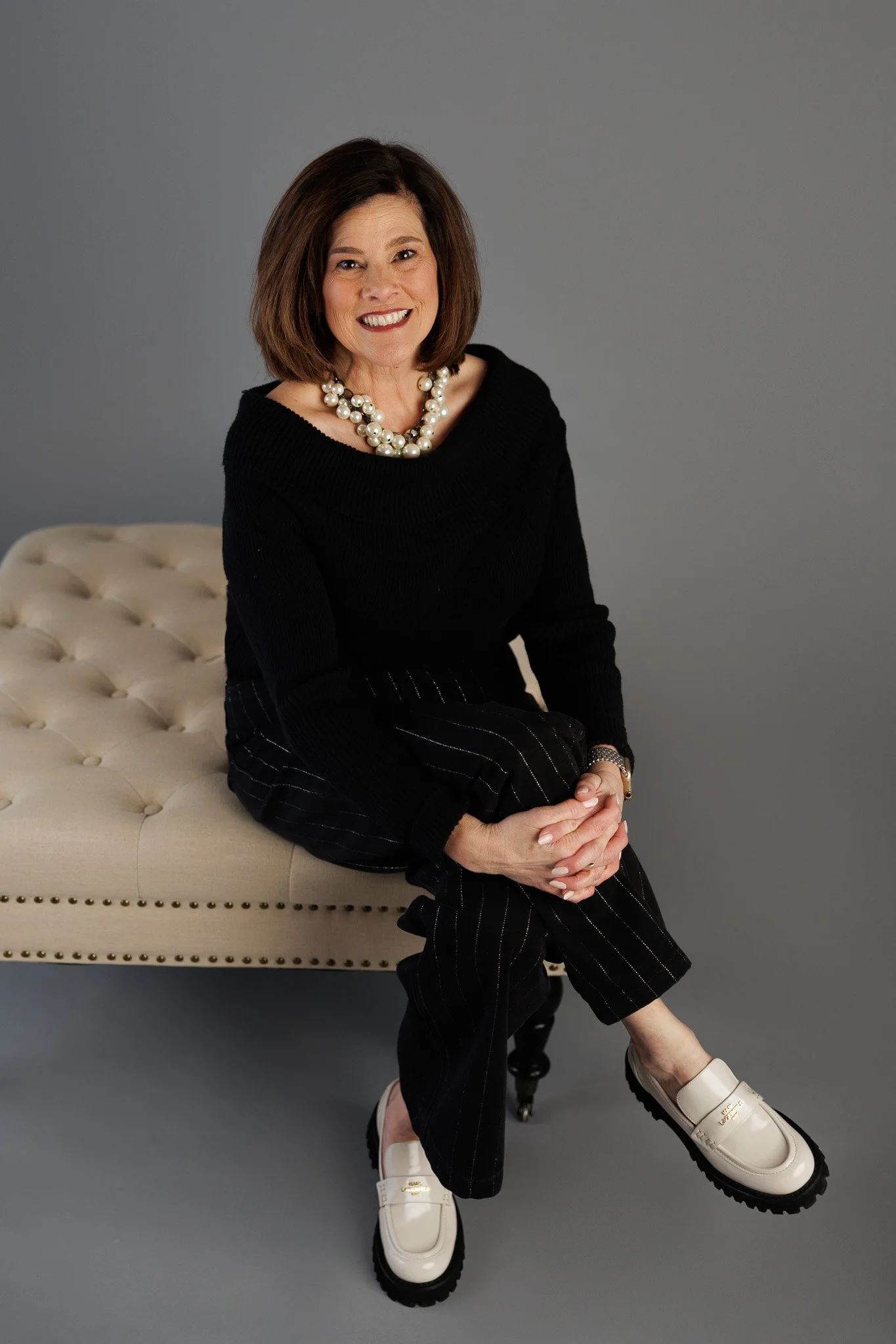 A woman sitting on a tufted beige bench with her hands clasped, wearing a black top, pinstriped pants, ivory loafers, a pearl necklace, and a watch, against a gray background.