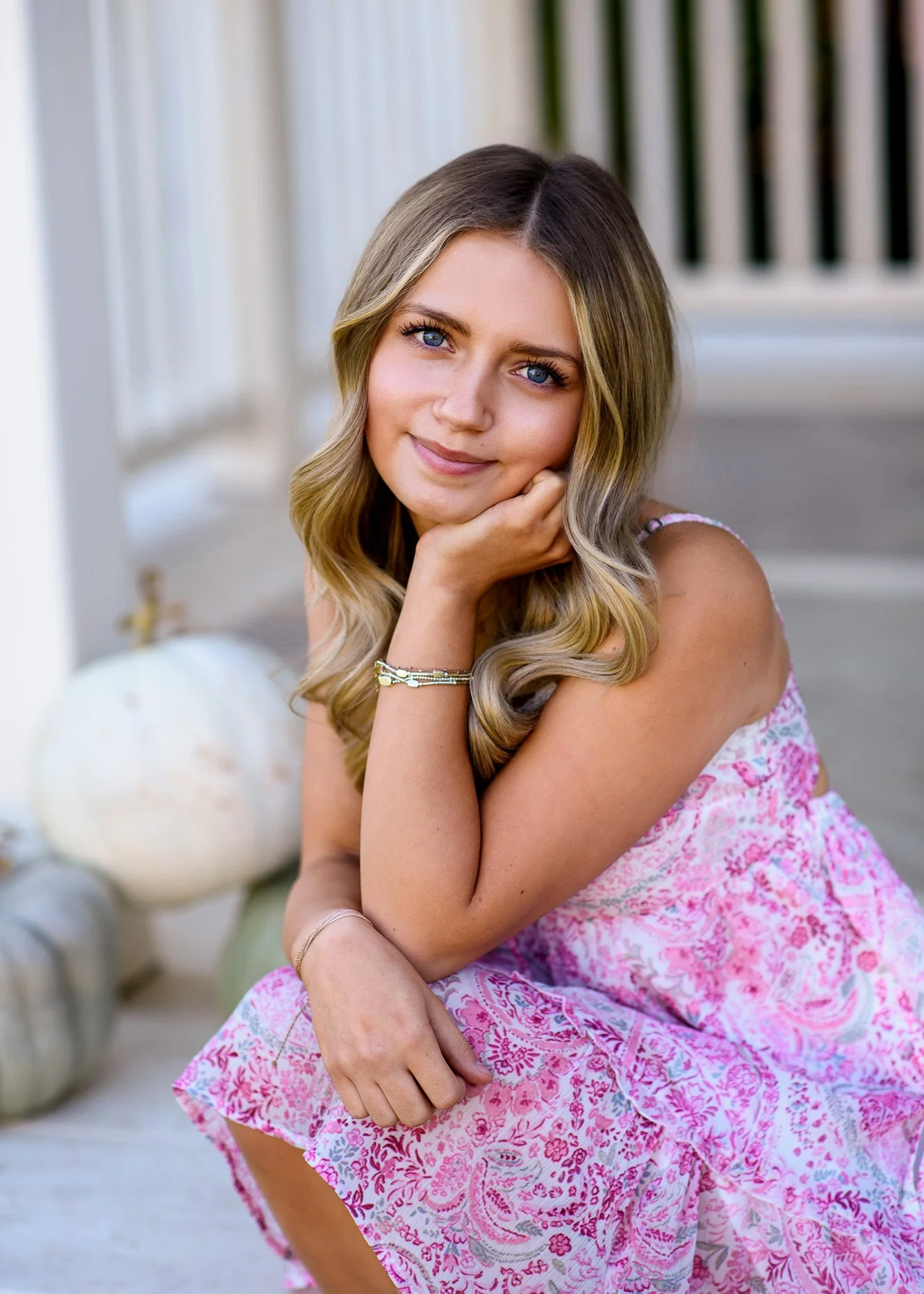 A young woman with blond, wavy hair sits outdoors, resting her chin on her hand and smiling gently at the camera, wearing a pink floral dress.