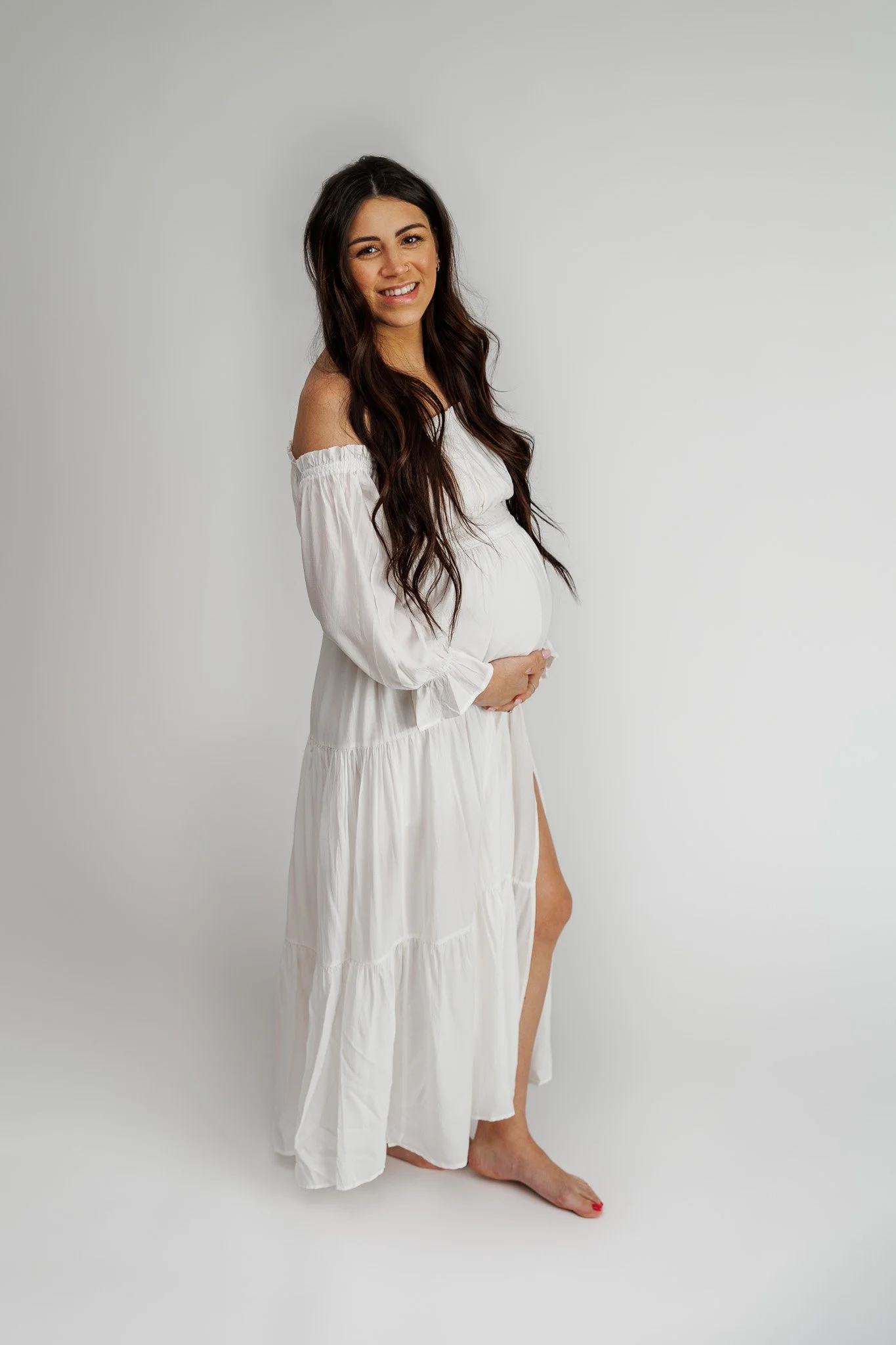 Pregnant woman in a white dress standing barefoot against a plain white background, smiling and gently holding her belly.