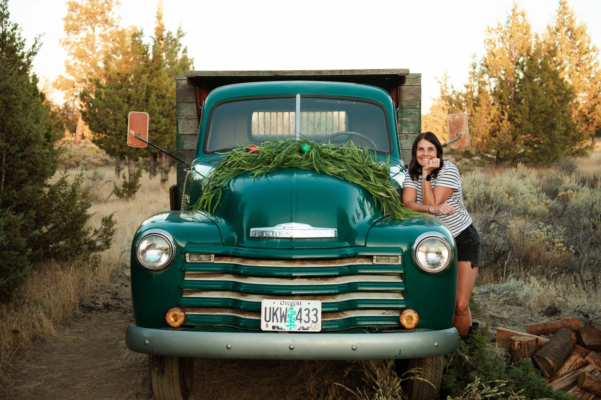 A smiling woman leaning on the front of a vintage green Chevrolet truck decorated with pine branches and Christmas ornaments, outdoors in a natural setting with trees and shrubs.