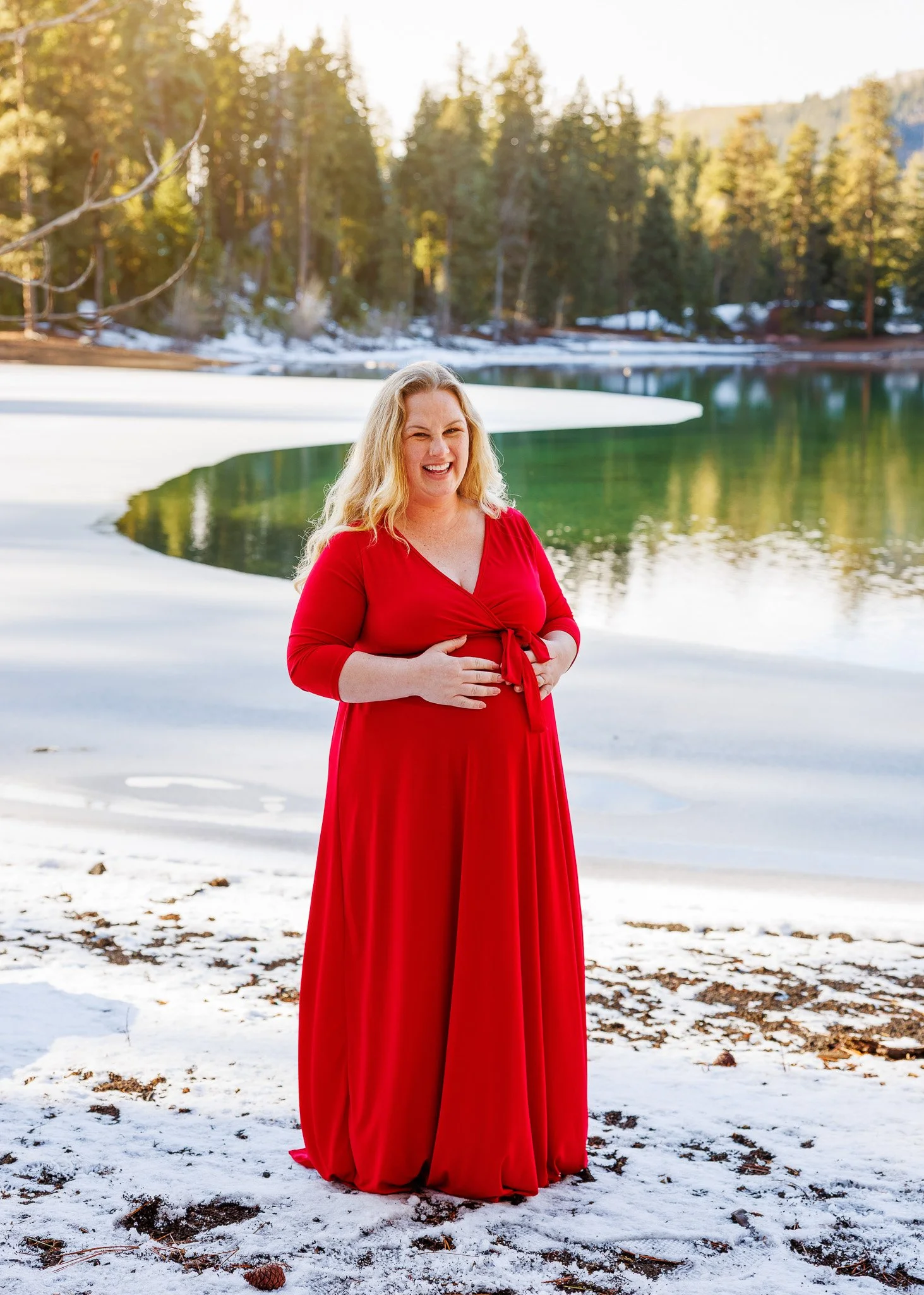 A woman in a long red dress standing on snow-covered ground near a partially frozen lake, with a forest of pine trees in the background, smiling and holding her pregnant belly.
