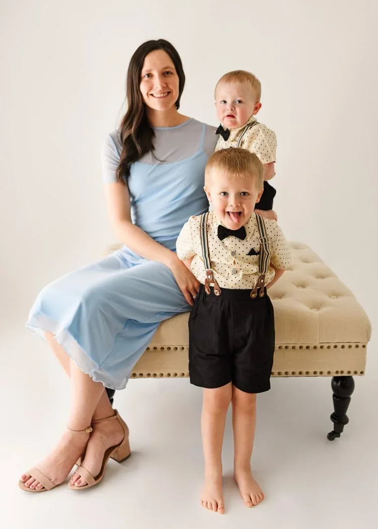 A woman with long dark hair, wearing a light blue dress and beige high heels, sitting on a beige upholstered bench. She has two young boys with light hair and fair skin, both dressed in matching outfits with polka-dotted shirts, bow ties, and suspend