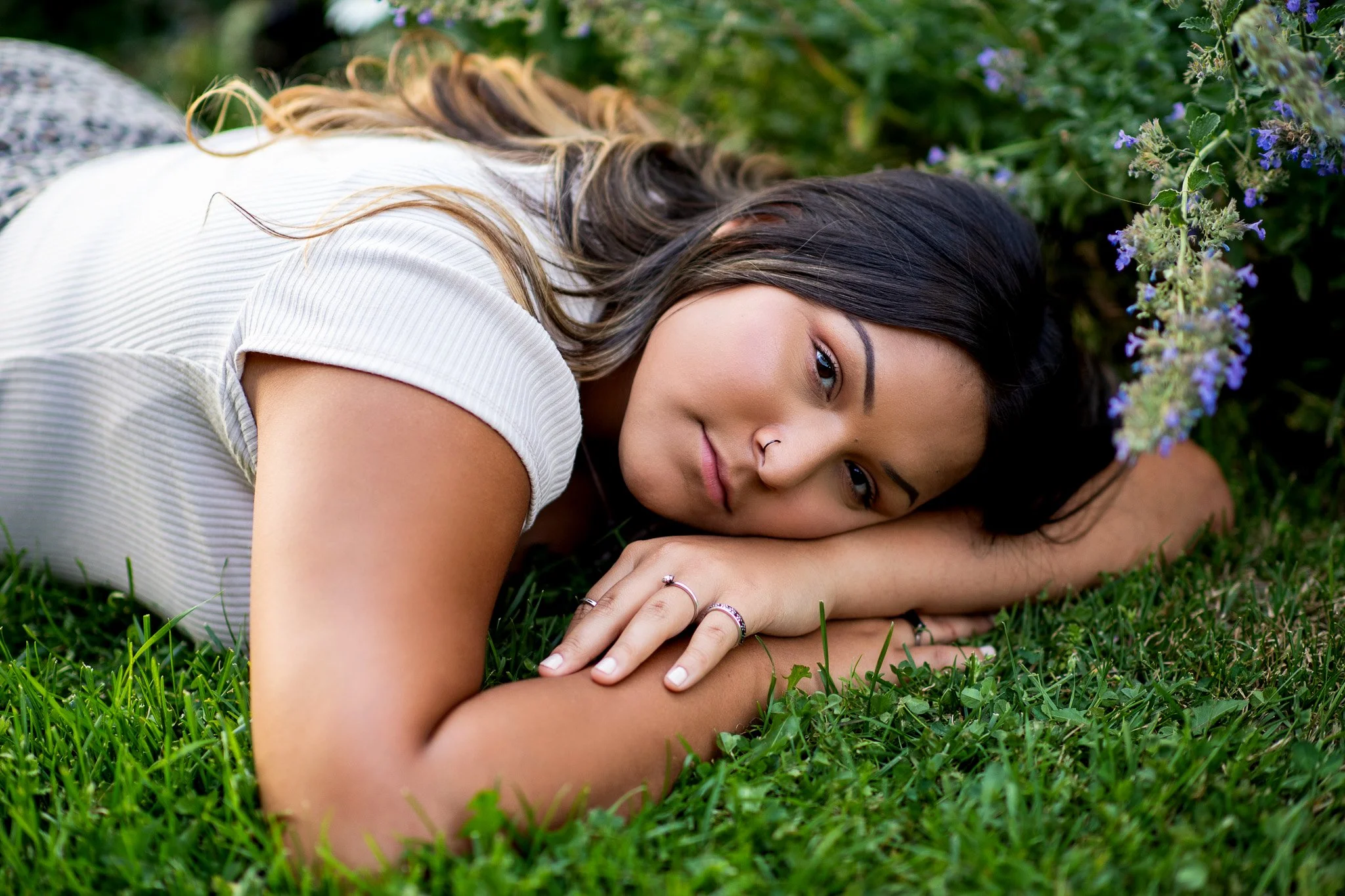 A young woman with long black hair lying on green grass next to purple flowers, looking at the camera with a gentle smile.
