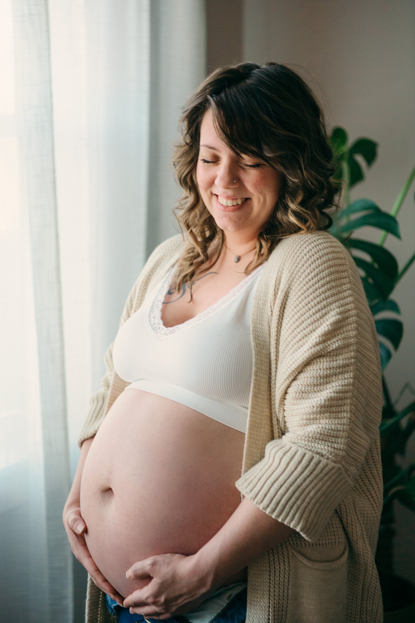 Smiling pregnant woman standing near a window with curtains, gently holding her pregnant belly with her hands.