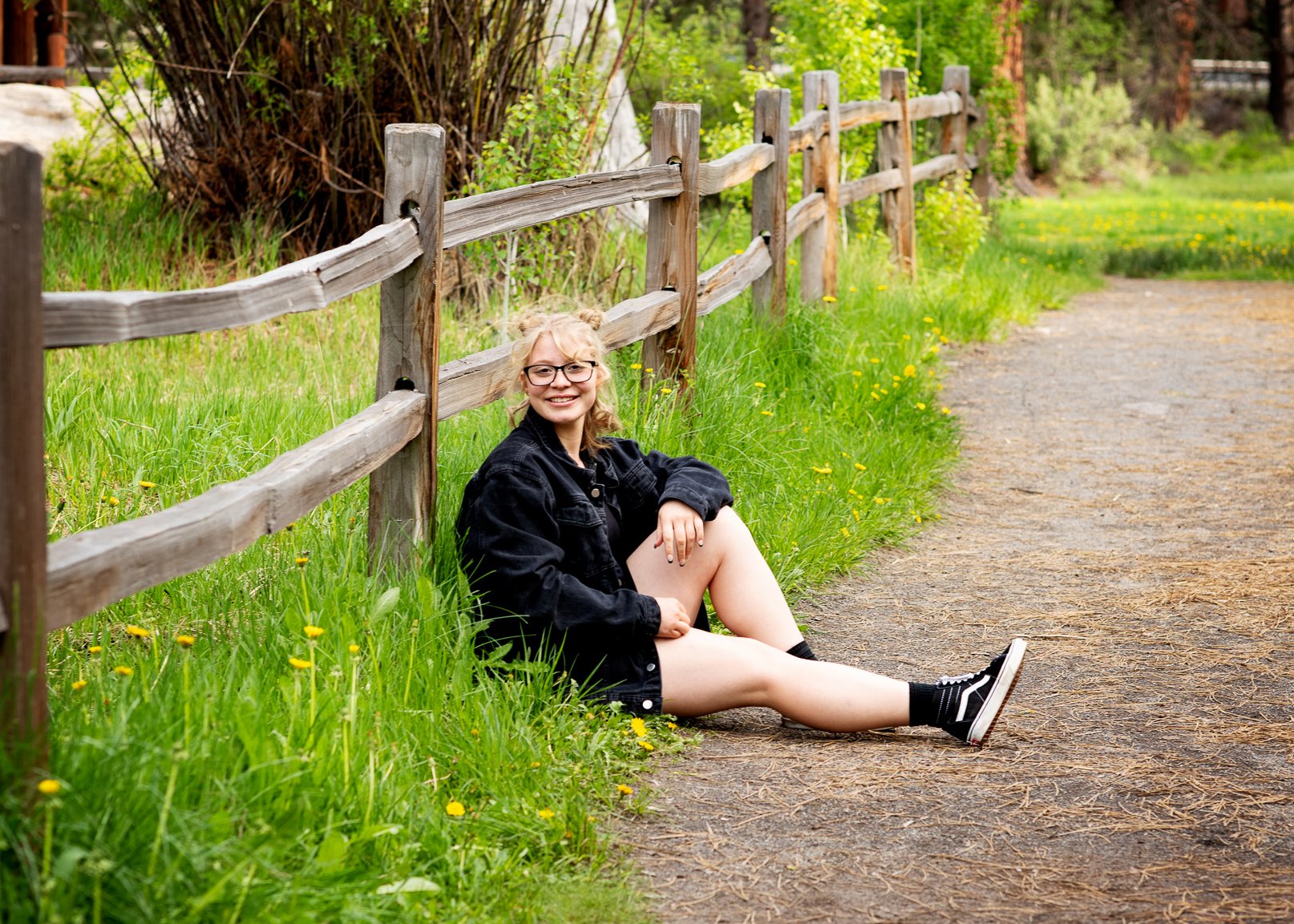 A young woman with curly blonde hair, glasses, wearing a black jacket, black shorts, and black sneakers, sitting on the grassy side of a dirt path beside a wooden fence in a green park, smiling at the camera.