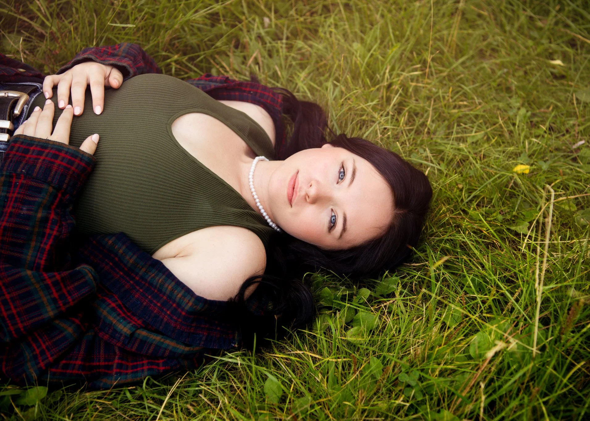 A woman with long dark hair, blue eyes, and wearing a pearl necklace, lies on her back on green grass, looking at the camera with a neutral expression. She is dressed in an olive green top with a cutout and a plaid shirt, with one hand resting on her