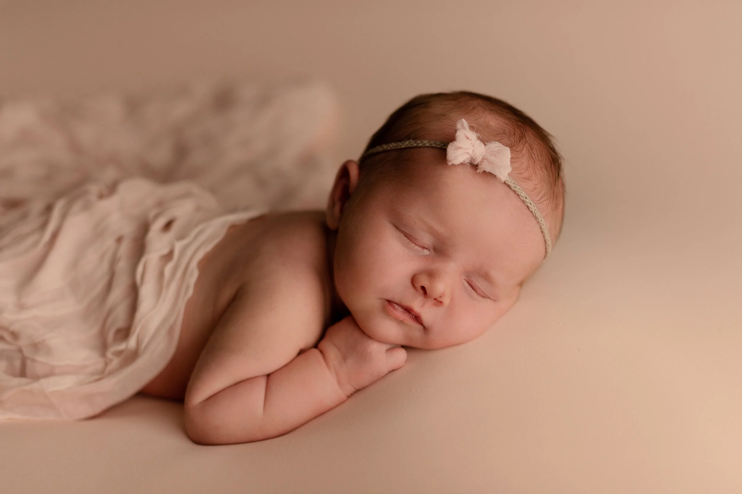 A newborn baby girl sleeping on her side with a beige headband and a small bow, resting her head on her hand, with a beige blanket partially covering her.