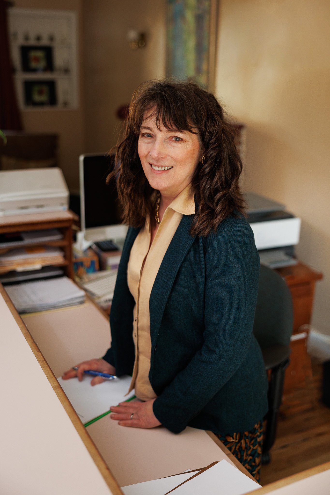 A woman with dark curly hair smiling, standing at an office desk with papers and a computer.